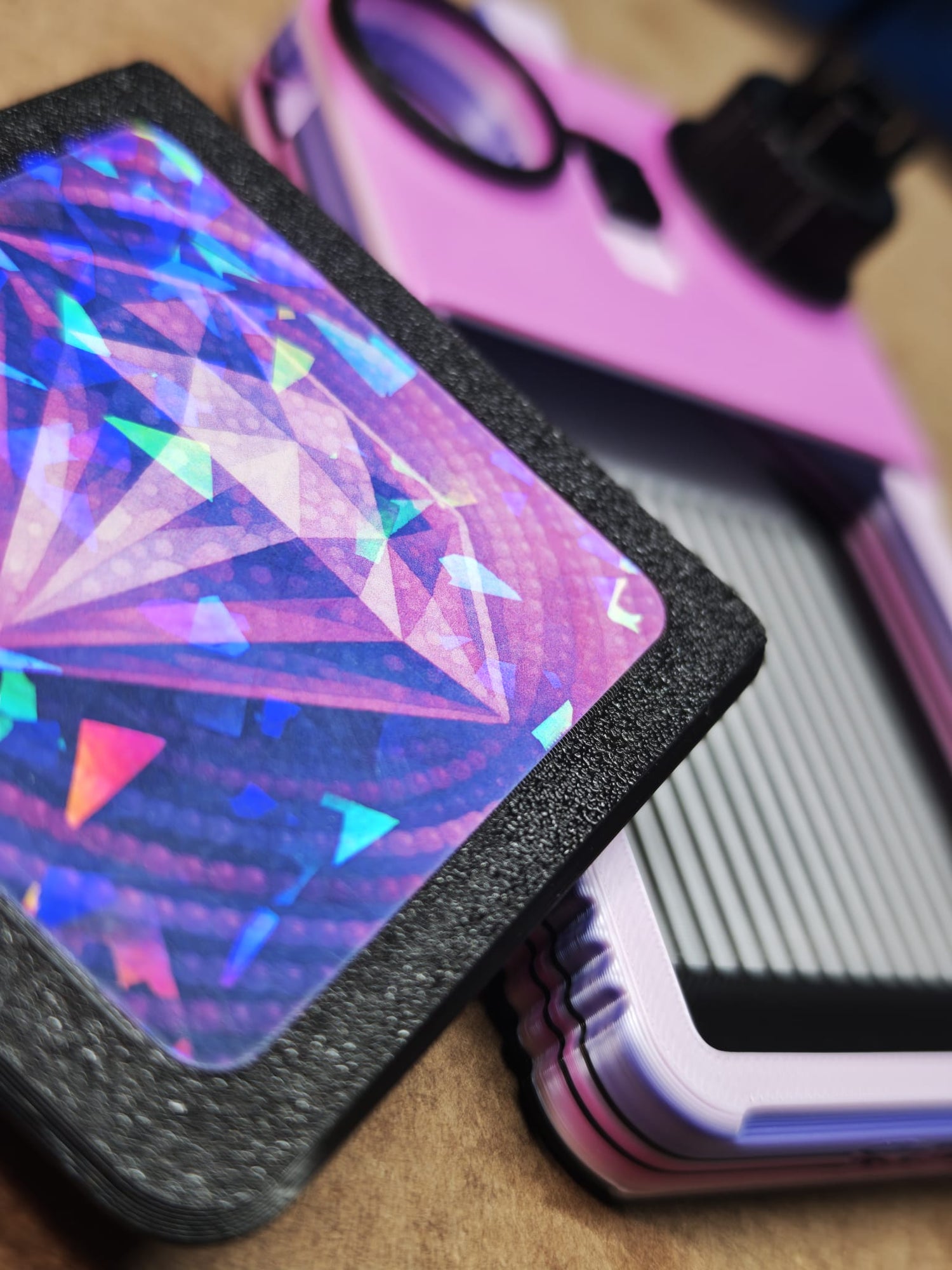 Eine Nahaufnahme einer leuchtenden, holografischen Karte mit Rautenmuster neben einem violett-schwarzen Objekt mit runder Linse, möglicherweise einer Kamera oder einer Handyhülle. Der Hintergrund ist unscharf.