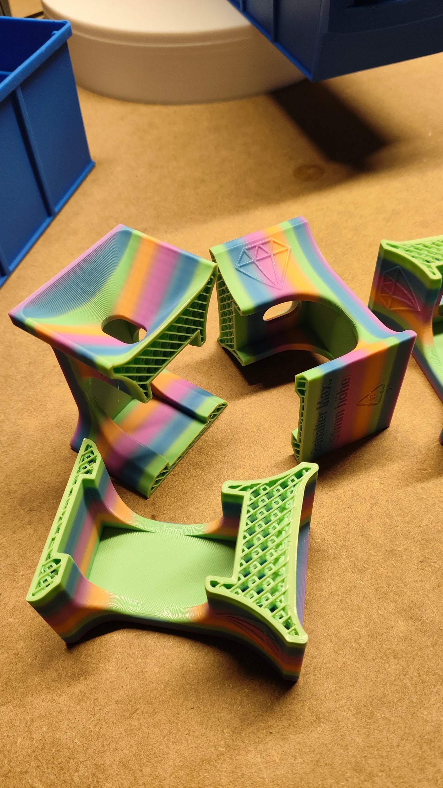 Die farbenfrohen 3D-gedruckten Objekte auf einer Holzoberfläche zeigen geometrische Designs mit offenen Flächen und Texturen. Mit gestreiften Regenbogen- und einfarbigen grünen Mustern ähneln diese Kreationen denen aus dem Set „Diamant-Malerei-Trichter für runde Dosen“ von New Plastics.