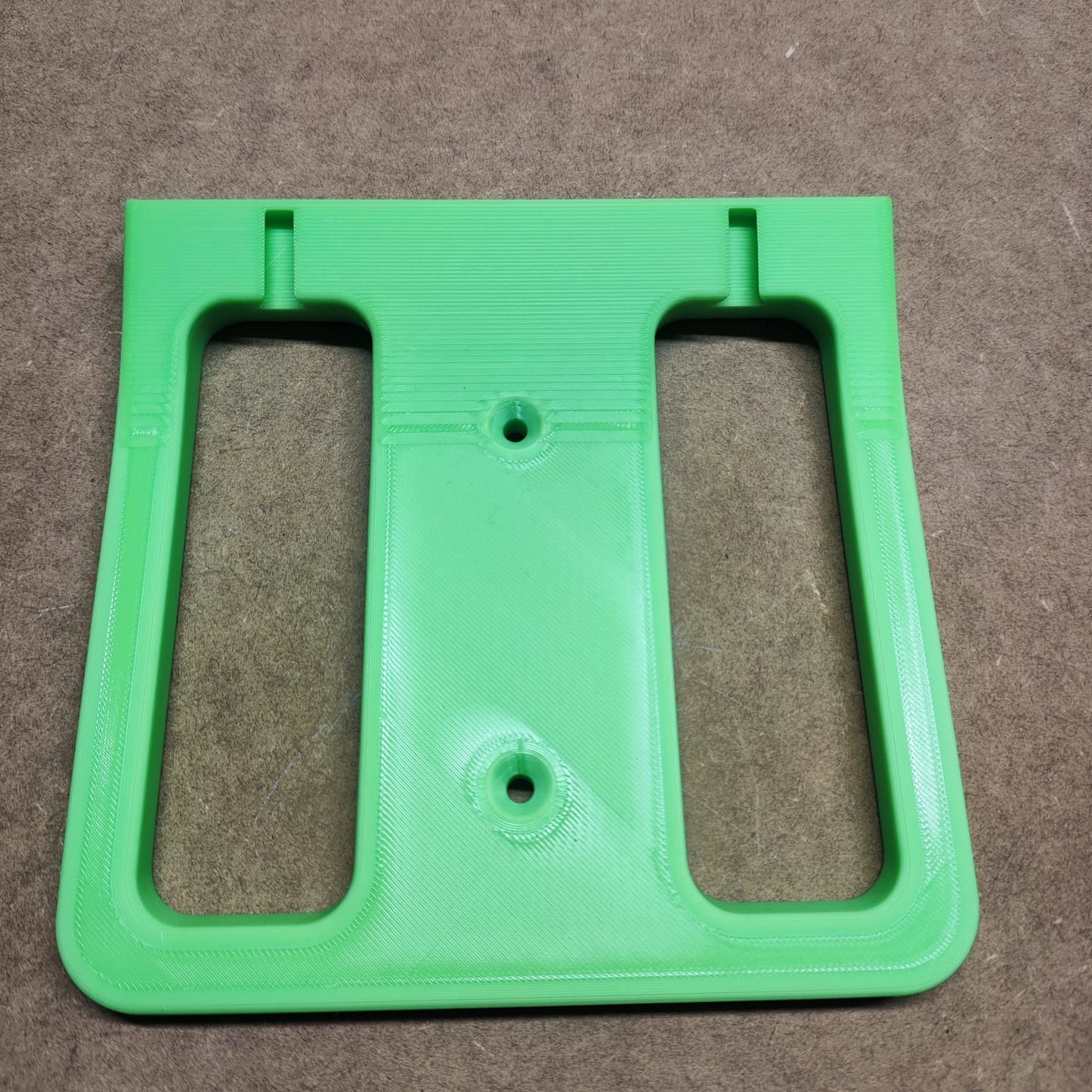 Eine grüne 3D-gedruckte Bodenplatte für Steinchen Garage von New Plastics mit zwei rechteckigen Ausschnitten ruht auf einer braunen strukturierten Oberfläche. Sie könnte Ihr Regalsystem ergänzen oder ein Diamond Painting-Regal perfekt unterstützen.