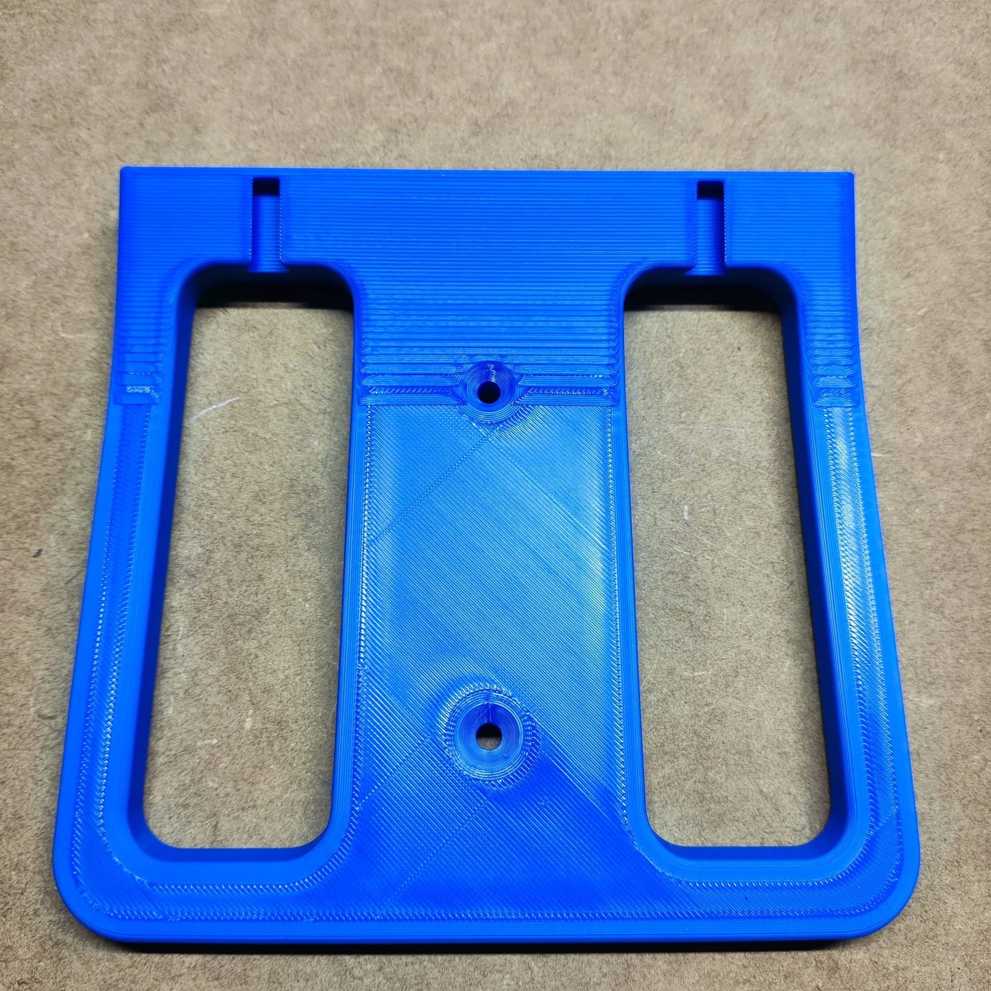Ein blaues 3D-gedrucktes Kunststoffbauteil von New Plastics, ähnlich einer Bodenplatte für Steinchen Garage, hat zwei rechteckige Öffnungen und zwei runde Löcher. Es bildet einen Kontrast zur braunen strukturierten Oberfläche, auf der es ruht.