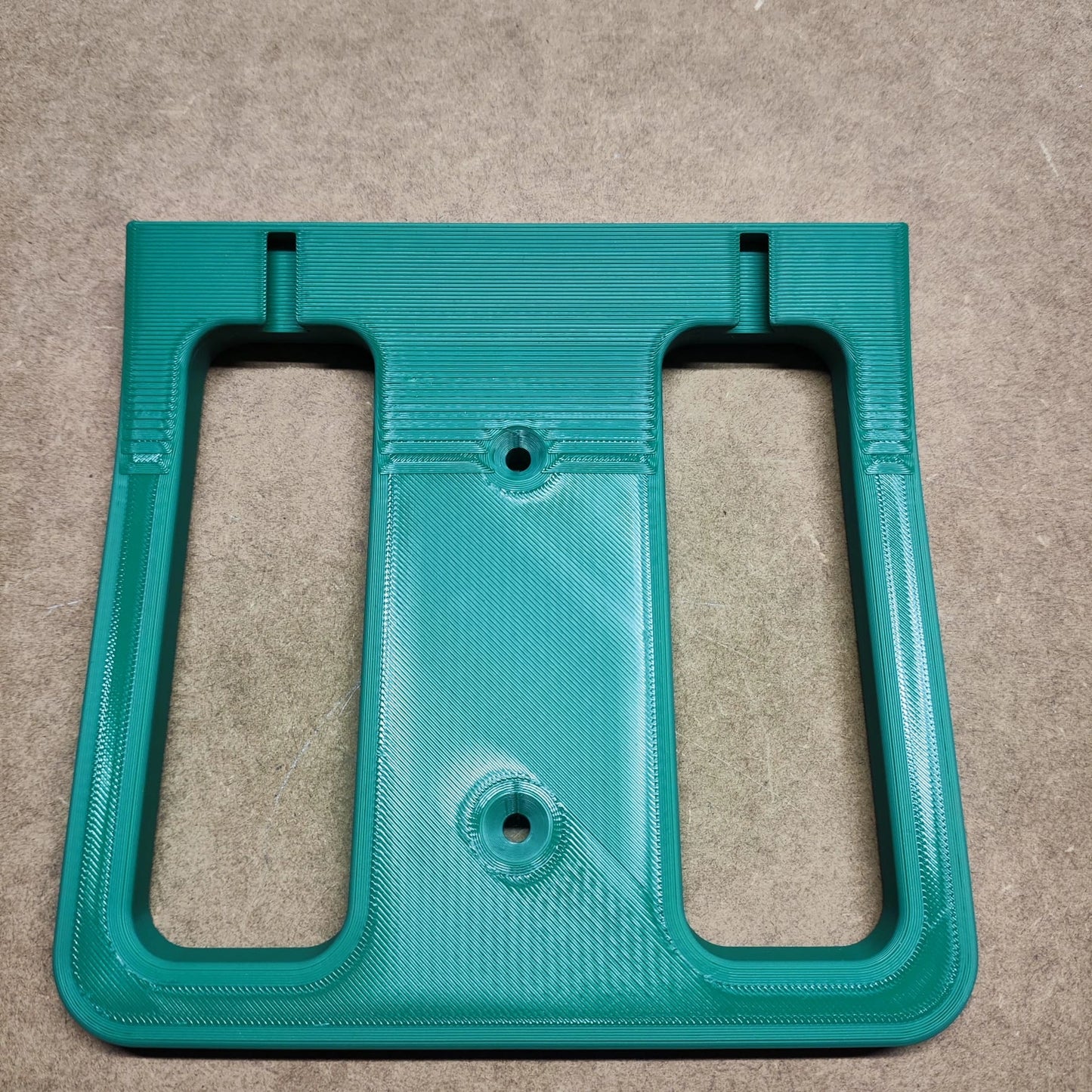Ein grünes 3D-gedrucktes New Plastics-Teil „Bodenplatte für Steinchen Garage“ mit zwei rechteckigen Ausschnitten und drei kreisrunden Löchern liegt auf einer braunen Oberfläche. Es könnte als Komponente für ein Regalsystem oder zur Verbesserung Ihrer Diamond Painting-Regal-Baugruppe verwendet werden.