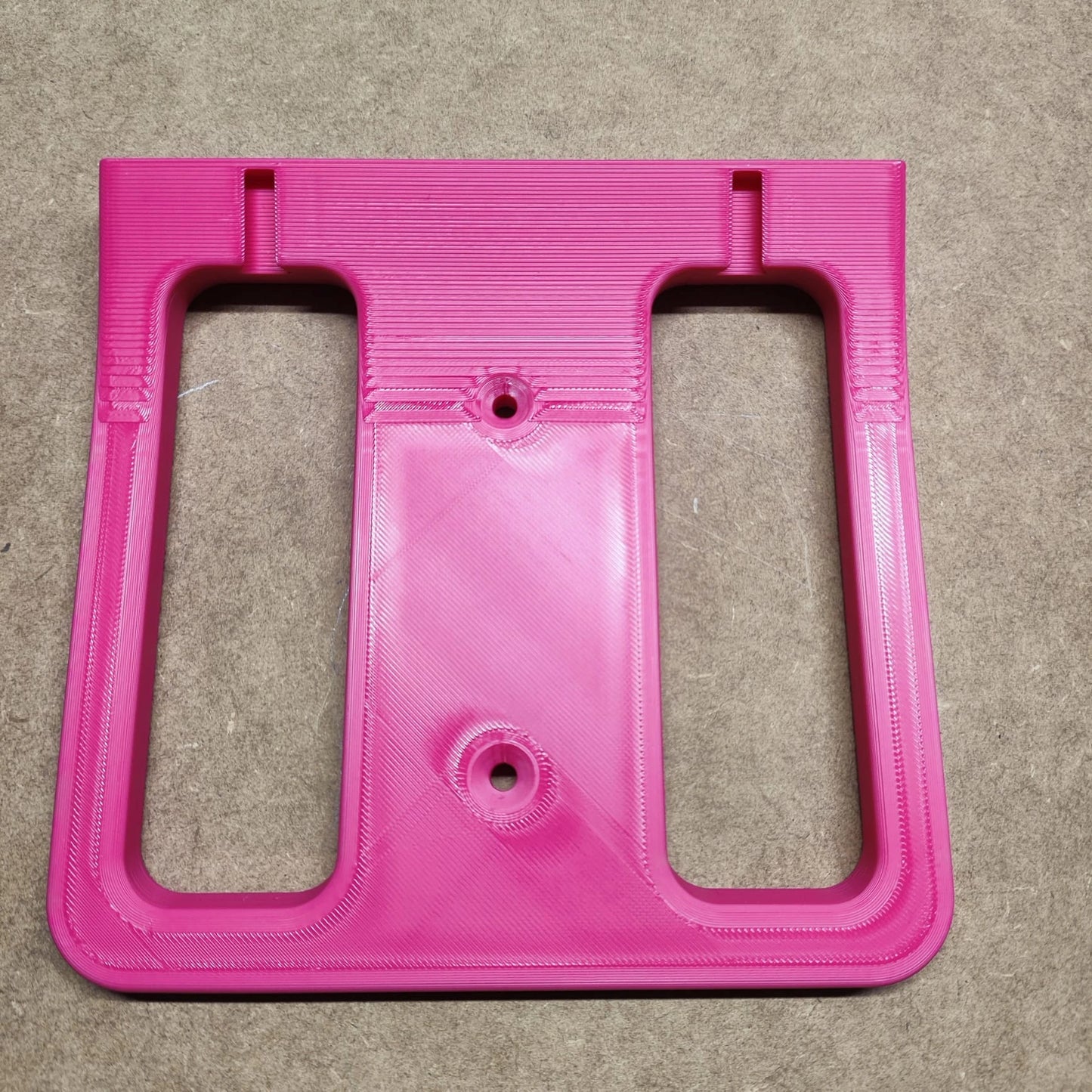 Eine leuchtend rosa, rechteckige, 3D-gedruckte Kunststoffhalterung aus der „Bodenplatte für Steinchen Garage“ von New Plastics mit zwei großen rechteckigen Ausschnitten und drei Schraubenlöchern ruht auf einer strukturierten beigen Oberfläche.
