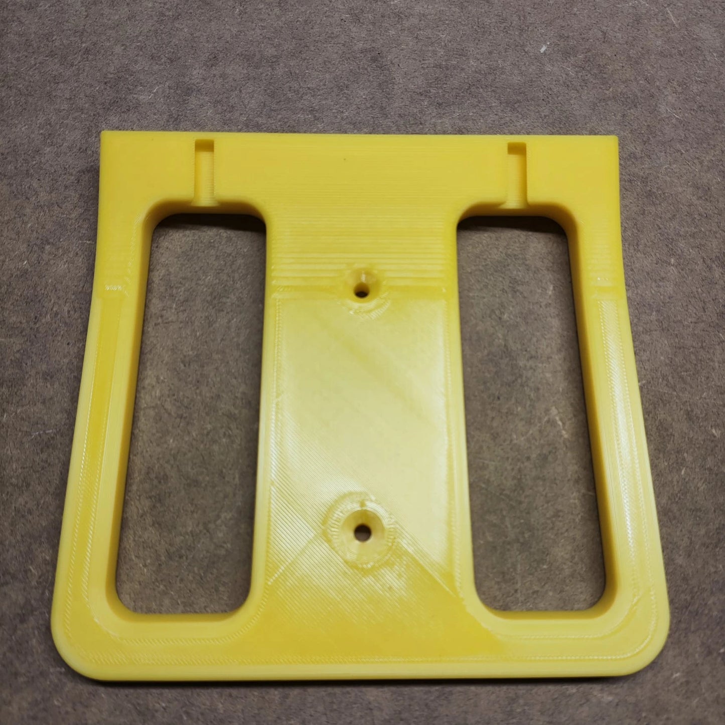 Die Bodenplatte für Steinchen Garage von New Plastics ist ein gelbes Kunststoffteil mit zwei rechteckigen Ausschnitten und drei Löchern, ideal für den Einsatz in einem Regalsystem oder als Bodenplatte. Sie ruht auf einem strukturierten braunen Hintergrund.