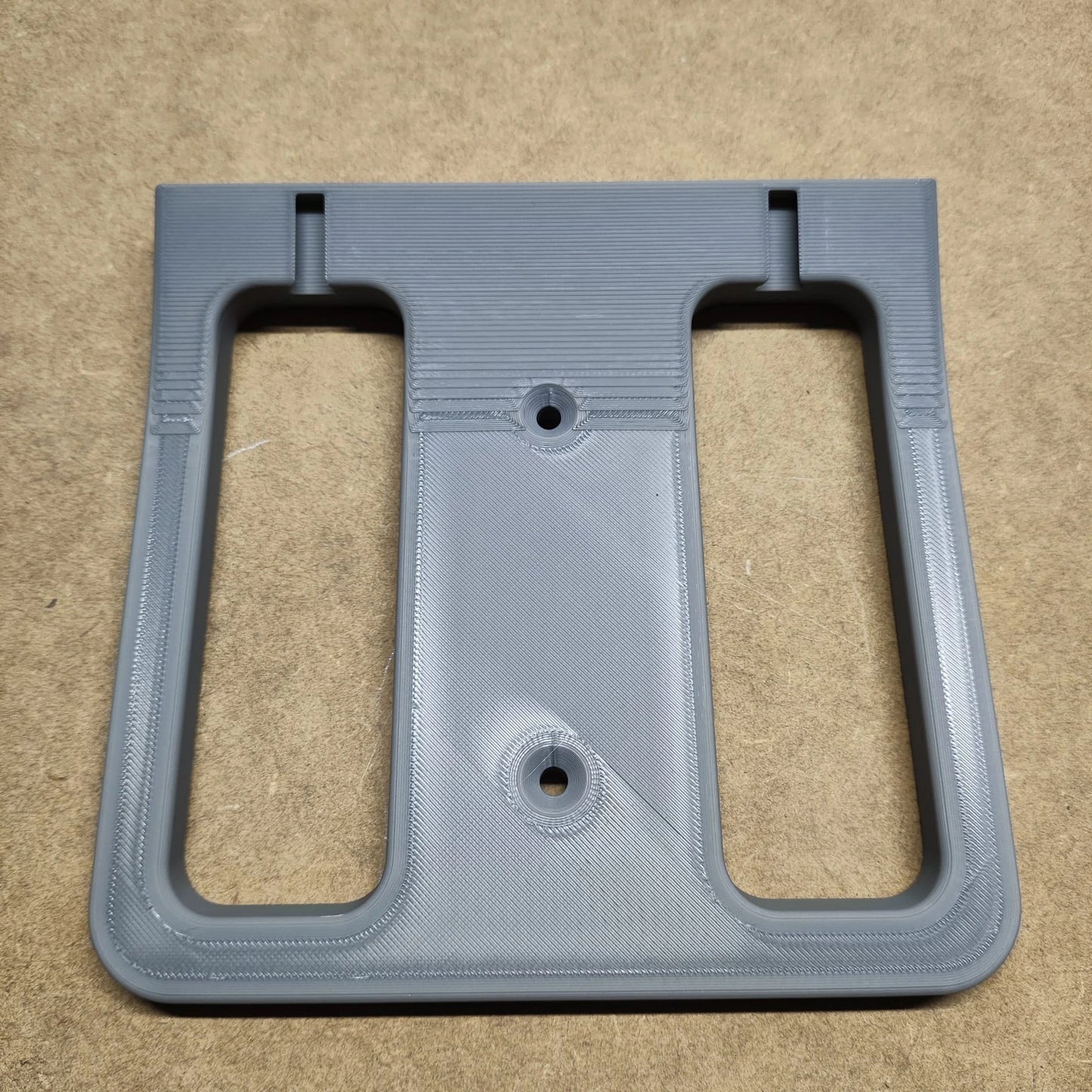 Ein graues, rechteckiges 3D-gedrucktes Objekt, das einer Bodenplatte für Steinchen Garage von New Plastics ähnelt und zwei große Ausschnitte und zwei kreisförmige Schraubenlöcher aufweist, ruht auf einer beigen Oberfläche.