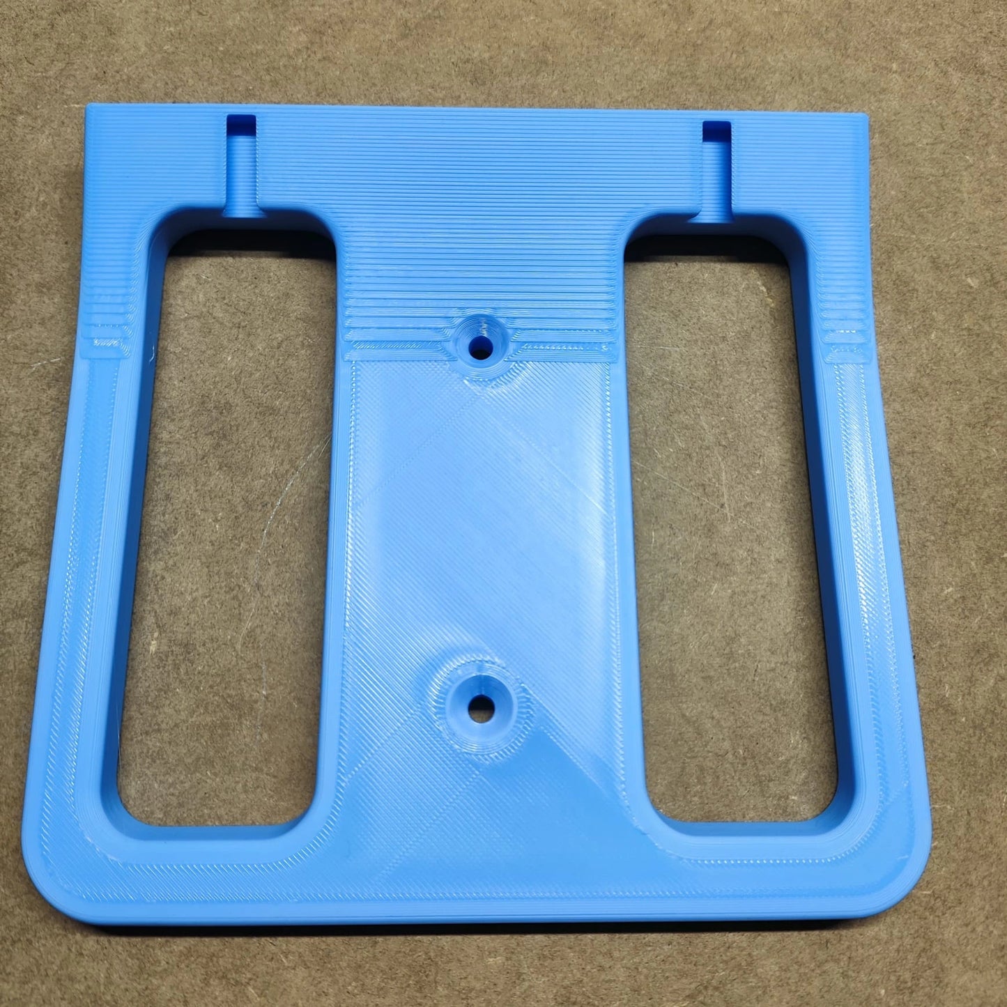 Ein blaues, rechteckiges Kunststoffbauteil aus der „Bodenplatte für Steinchen Garage“ von New Plastics weist zwei große Ausschnitte auf. Es hat eine glatte Oberfläche mit 3D-Drucklinien und drei Schraubenlöchern – zwei oben, eines unten in der Mitte – ideal zur Ergänzung eines Regalsystem-Designs auf einer strukturierten braunen Oberfläche.