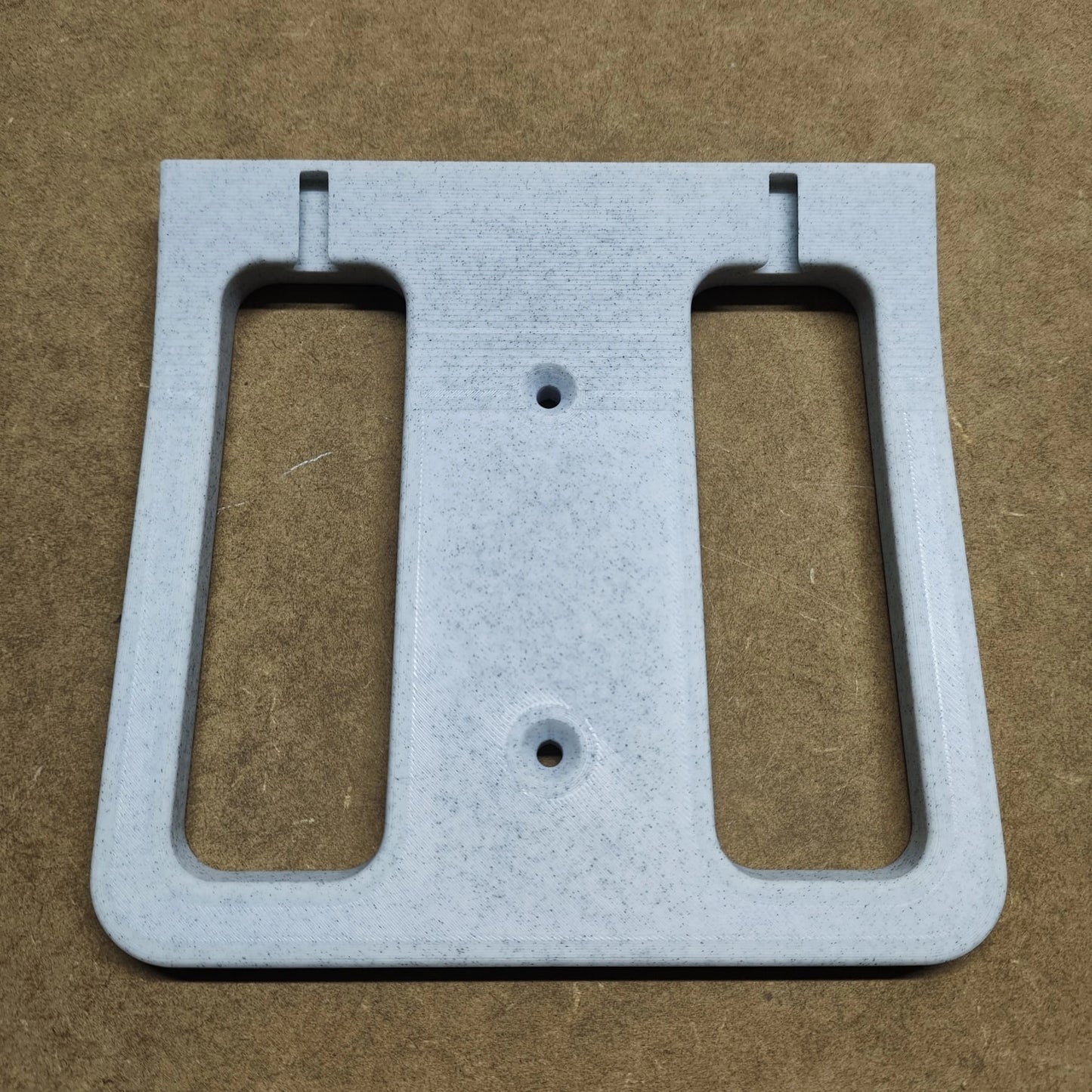 Die New Plastics Bodenplatte für Steinchen Garage, eine graue, rechteckige Halterung mit zwei seitlichen Schlitzen und zentralen Schraubenlöchern, ziert eine braun strukturierte Oberfläche.