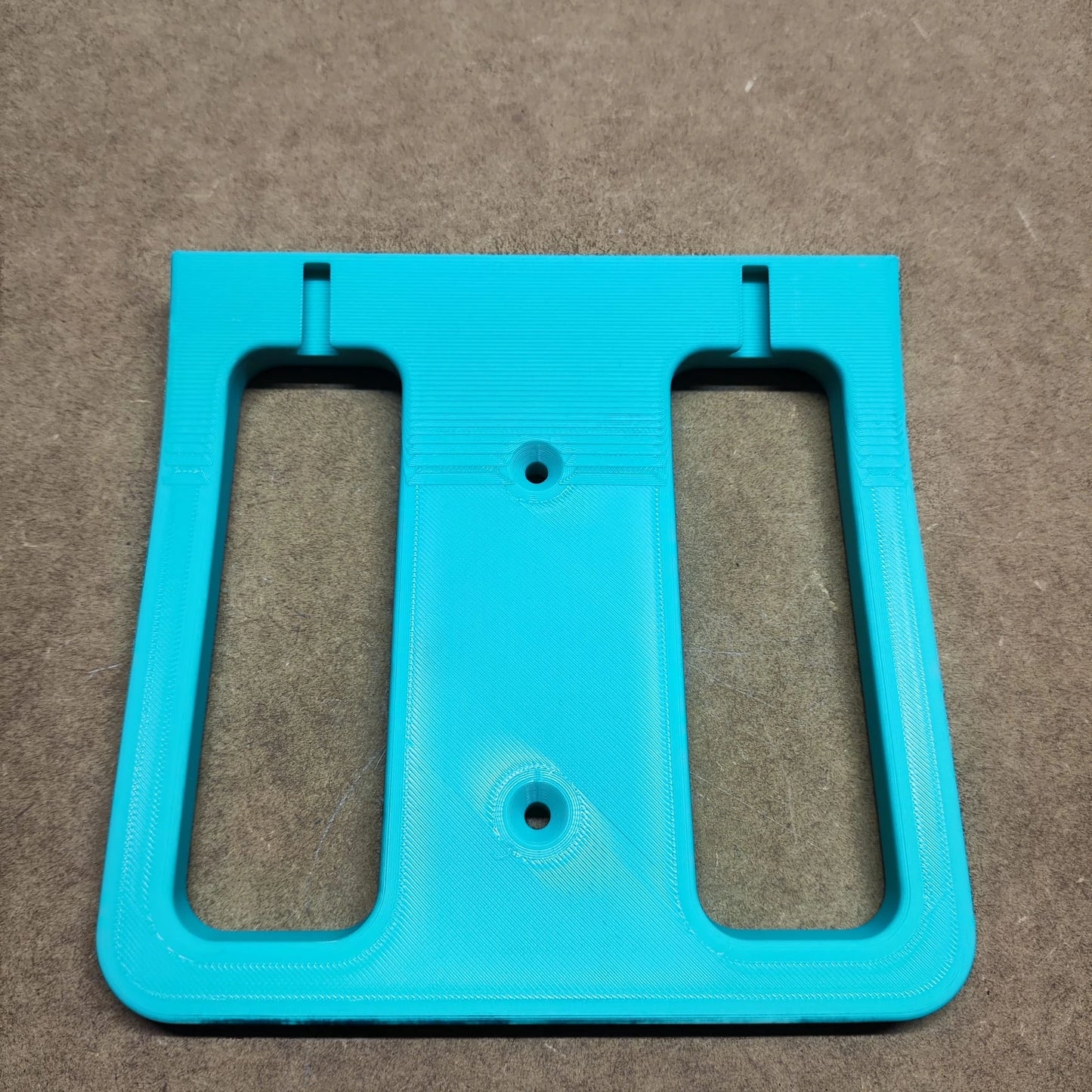 Eine türkisfarbene 3D-gedruckte „Bodenplatte für Steinchen Garage“ von New Plastics mit einer rechteckigen Basis und zwei vertikalen Schlitzen wie ein Miniatur-Regalsystem ruht auf einer braunen strukturierten Oberfläche.