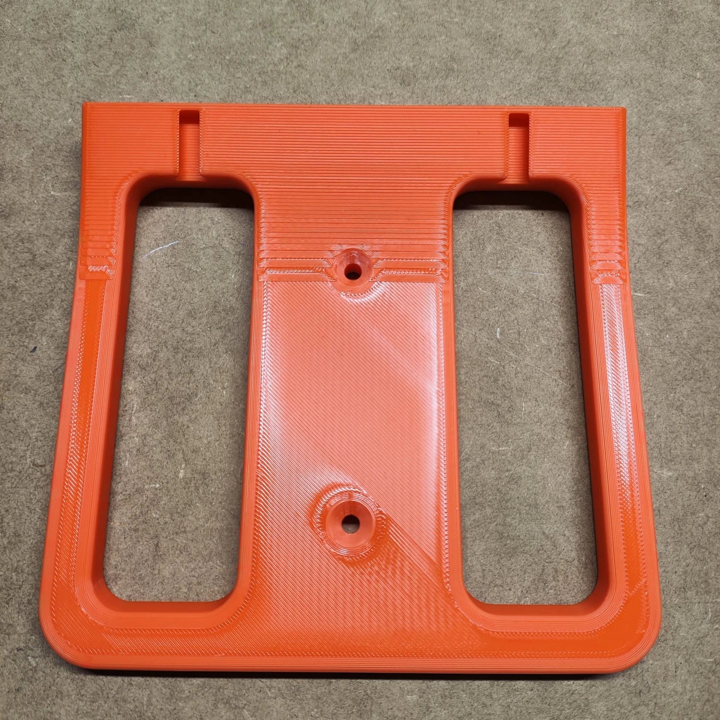 Das 3D-gedruckte orangefarbene Kunststoffteil von New Plastics ähnelt einer Bodenplatte für Steinchen Garage, hat eine rechteckige Basis, zwei vertikale Ausschnitte und eine strukturierte Oberfläche. Es ist ideal für jedes Regalsystem oder Diamond Painting-Regal-Setup und ruht auf einem braunen Hintergrund.