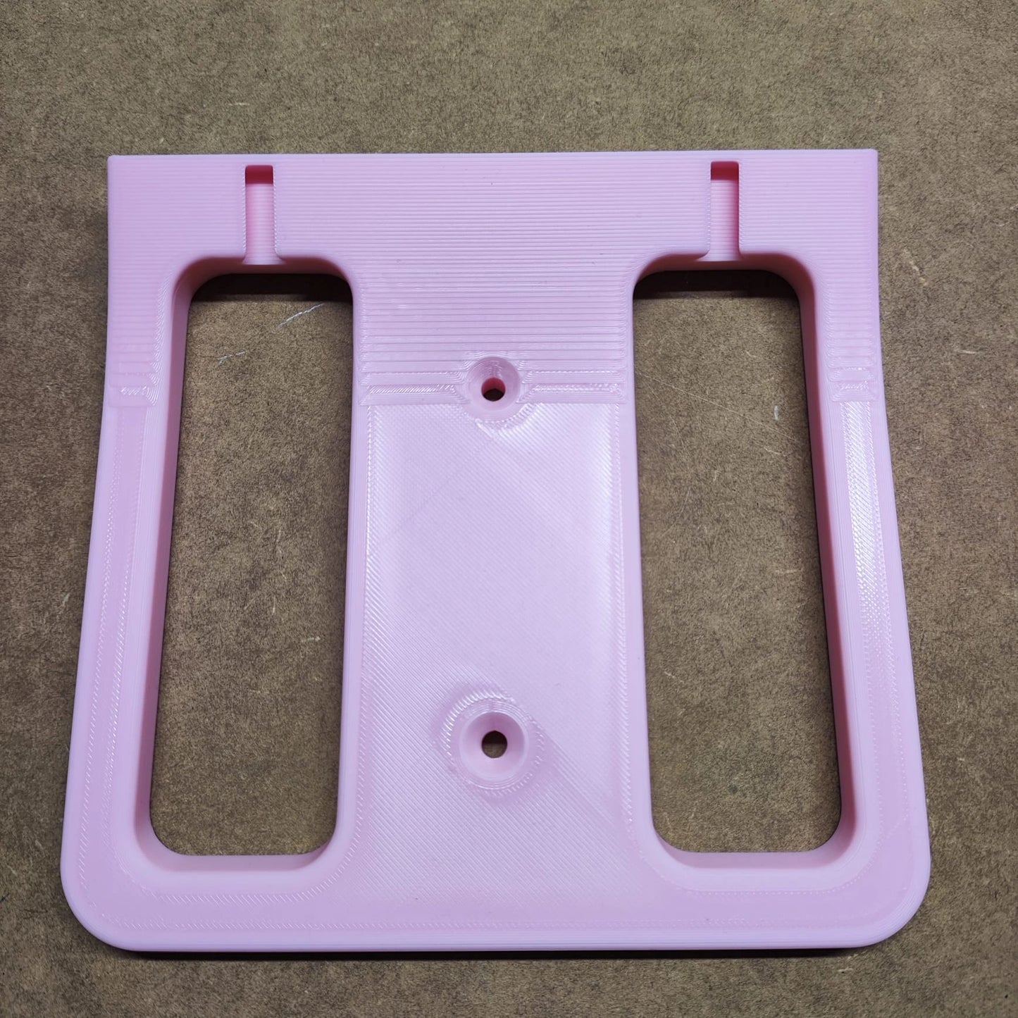 Ein rosa 3D-gedrucktes Objekt, das einem miniaturisierten Regalsystem ähnelt, mit zwei vertikalen Ausschnitten und kreisförmigen Löchern in der Mitte, liegt auf einer braunen Oberfläche. Das strukturierte Stück ist die Bodenplatte für Steinchen Garage von New Plastics.