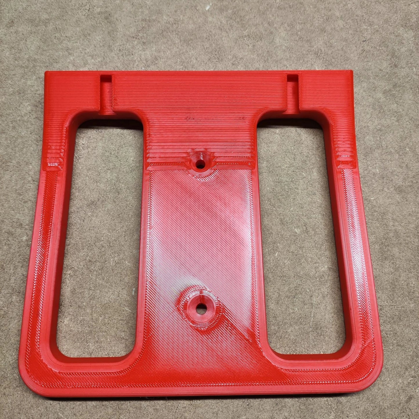 Eine 3D-gedruckte, rote, rechteckige New Plastics Bodenplatte für Steinchen Garage mit zwei Aussparungen und abgerundeten Kanten liegt auf einem braunen Strukturuntergrund. Sie verfügt über zwei kleine kreisrunde Löcher und ein größeres Loch in der Mitte zur Befestigung, perfekt für den Einsatz in einem Diamond Painting-Regal.