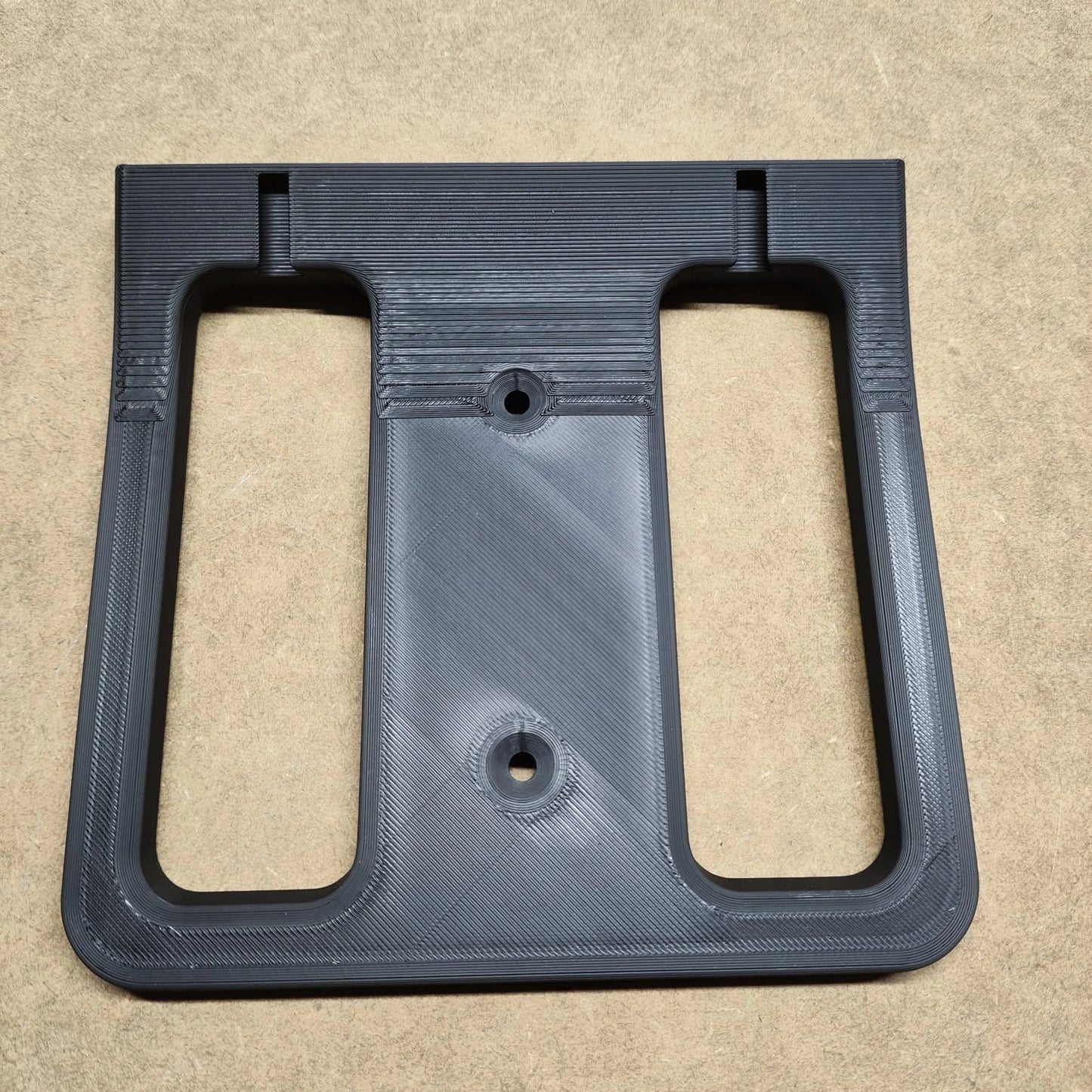 Ein schwarzer, rechteckiger Kunststoffrahmen von New Plastics, der an eine Bodenplatte für Steinchen Garage erinnert, weist zwei große Aussparungen und drei Schraubenlöcher auf. Er ruht auf einer beigen Oberfläche mit einer sichtbaren gerippten Textur für zusätzliche Designprägnanz.