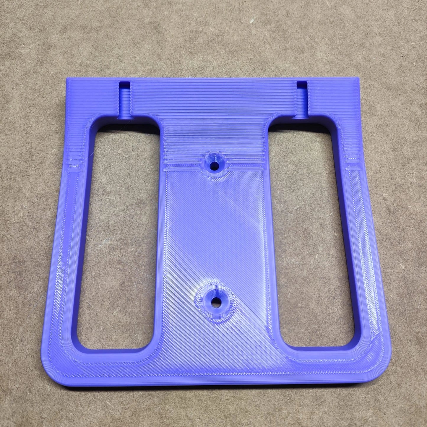 Eine violette, 3D-gedruckte rechteckige „Bodenplatte für Steinchen Garage“ von New Plastics mit zwei großen Ausschnitten und drei kleinen Löchern liegt auf einer braunen Oberfläche. Sie hat eine glatte Oberfläche mit sichtbaren Schichtmustern und ähnelt einem Teil eines komplizierten Regalsystems.