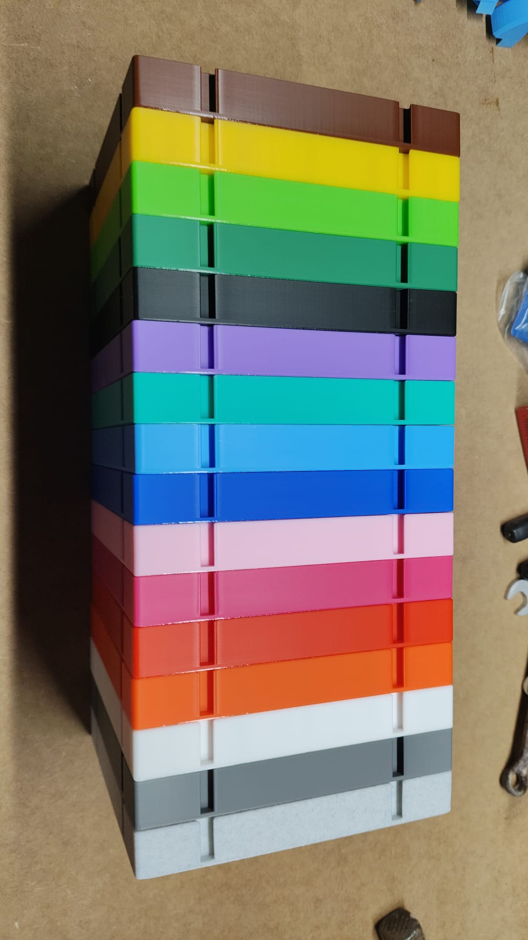 Die Steinchen-Garage Modulplatte für 25 Farbdosen Rund von New Plastics ähnelt mit ihrem vertikalen Stapel aus rechteckigen Blöcken in Grün, Gelb, Blau, Lila, Rosa, Rot und Grau auf einer hellbraunen Oberfläche einem lebendigen Diamantgemälde.