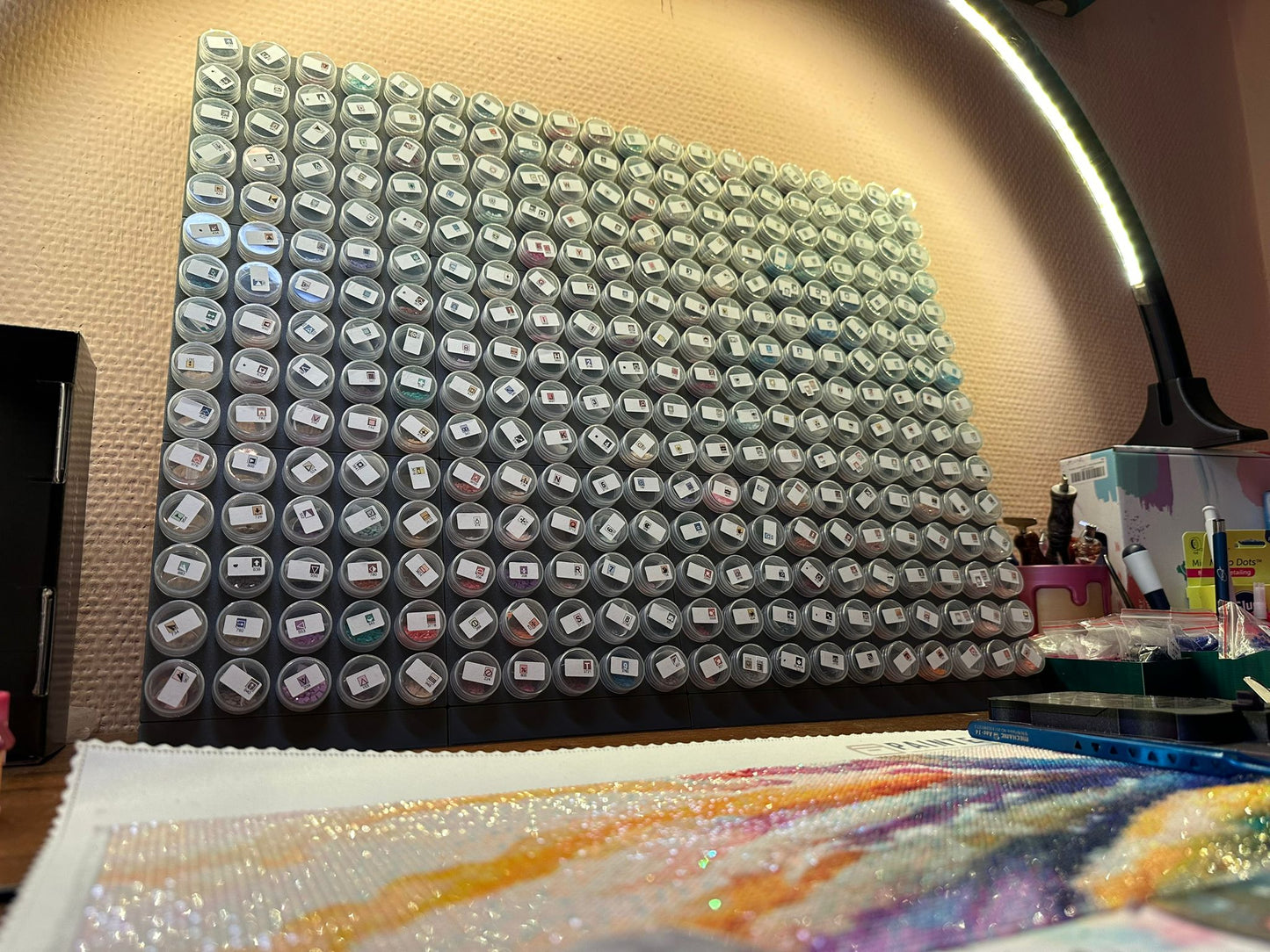 Eine Steinchen-Garage Modulplatte für 25 Farbdosen Rund von New Plastics zeigt zahlreiche kleine, beschriftete Behälter in einem Raster. Darunter ist ein farbenfrohes, halbfertiges Diamantgemälde mit Perlen und Zubehör zu sehen. Eine Deckenlampe beleuchtet den künstlerischen Arbeitsbereich.