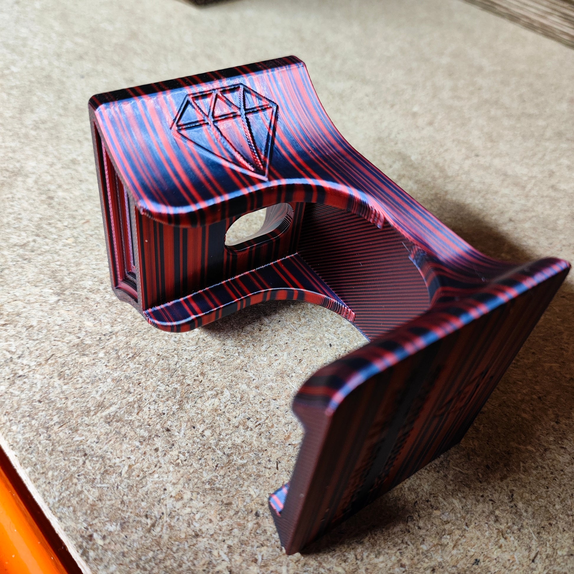 Ein 3D-gedrucktes Objekt aus dem Diamond Painting Trichter für runde Dosen von New Plastics weist ein rot-blaues Streifenmuster mit Ausschnitten und geometrischen Rautenformen auf. Es ruht auf einer strukturierten Oberfläche und zeigt sein geschwungenes, strukturiertes Design – möglicherweise zum sauberen Befüllen runder Dosen.
