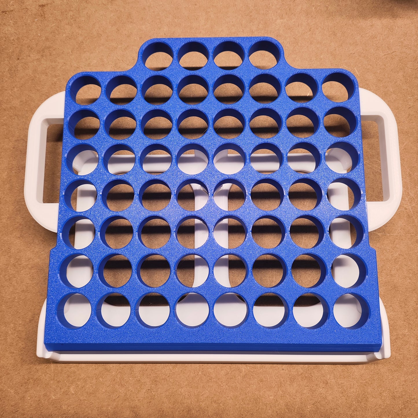 Eine kreisförmige Gitterplatte aus blauem Kunststoff von New Plastics mit mehreren Löchern ist auf einem weißen rechteckigen Rahmen mit Griffen wie bei einer Klappbar 60er von Steinchen-Garage angebracht, der auf einer braunen Oberfläche platziert ist.