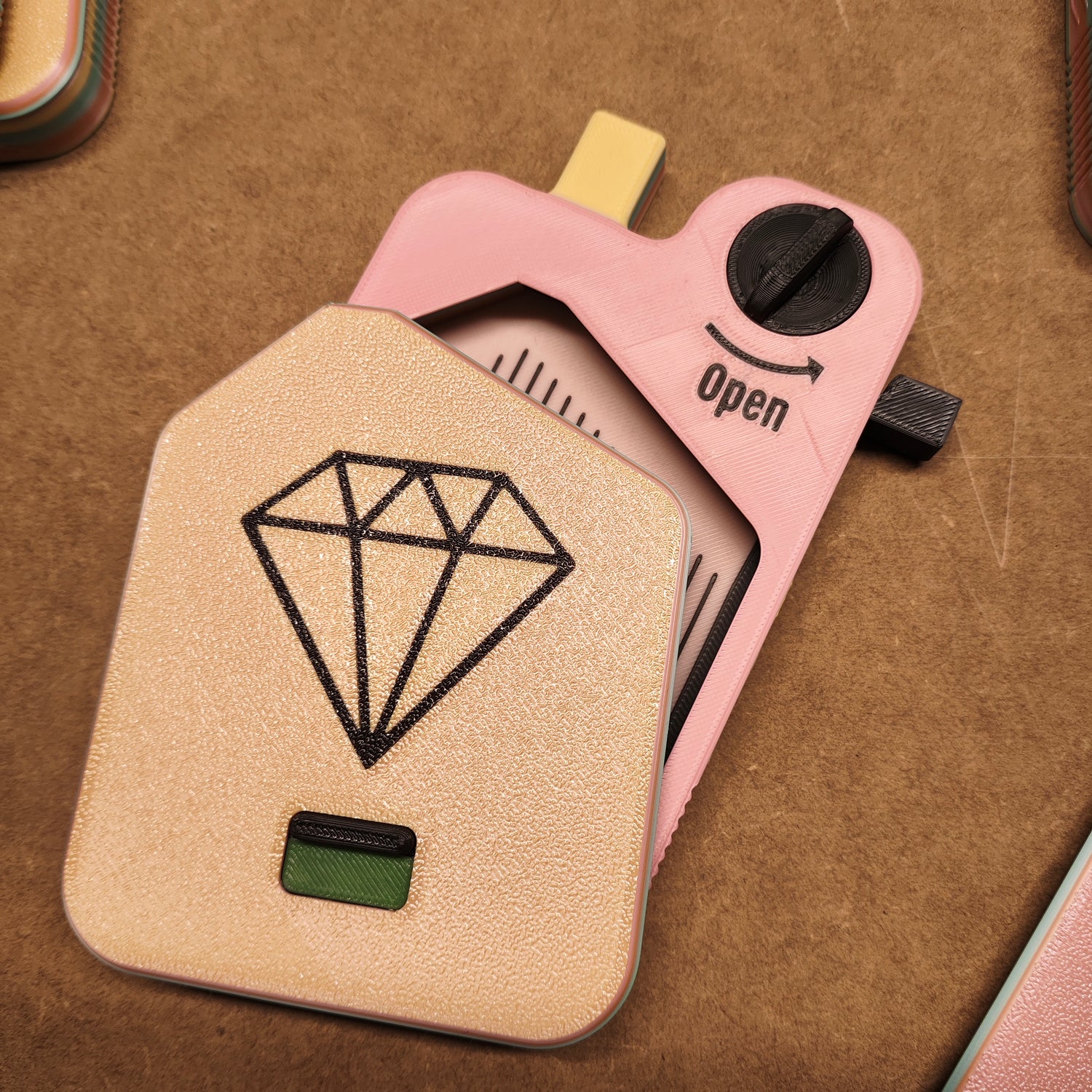 Eine Nahaufnahme des Diamond Painting Schiffchen S2 von New Plastics zeigt einen rosa Kunststoffkörper mit einer beigen Abdeckung mit einem schwarzen Rautensymbol. Ein Knopf mit der Aufschrift „Open“ zeigt den sicheren Verschlussmechanismus an, ein Pfeil zeigt die Richtung an, alles vor einem strukturierten braunen Hintergrund.