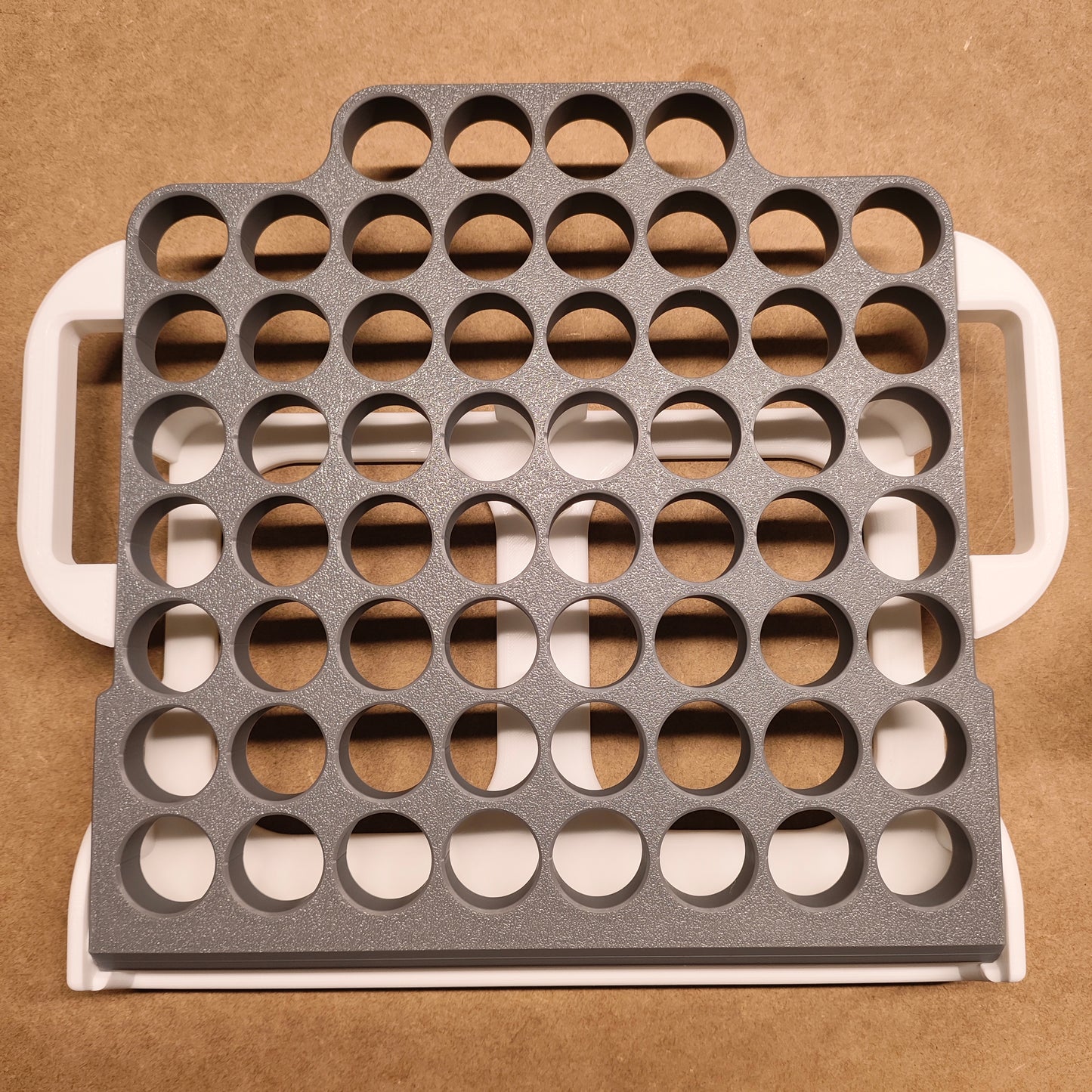 Eine graue Kunststoffschale mit Reihen kreisförmiger Löcher liegt auf einer beigen Oberfläche und dient als ideale New Plastics Steinchen-Garage Klappbar 60er Individuelle Farbgestaltung. Mit seinen weißen Griffen ist dieser Organizer perfekt für Bastel- oder Diamantmalerei-Enthusiasten, die ein effizientes Aufbewahrungssystem suchen.