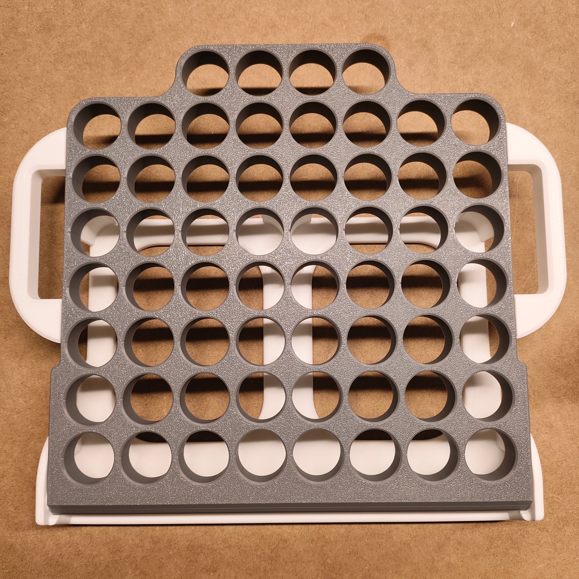 Eine graue Kunststoffschale mit Reihen kreisförmiger Löcher liegt auf einer beigen Oberfläche und dient als ideale New Plastics Steinchen-Garage Klappbar 60er Individuelle Farbgestaltung. Mit seinen weißen Griffen ist dieser Organizer perfekt für Bastel- oder Diamantmalerei-Enthusiasten, die ein effizientes Aufbewahrungssystem suchen.