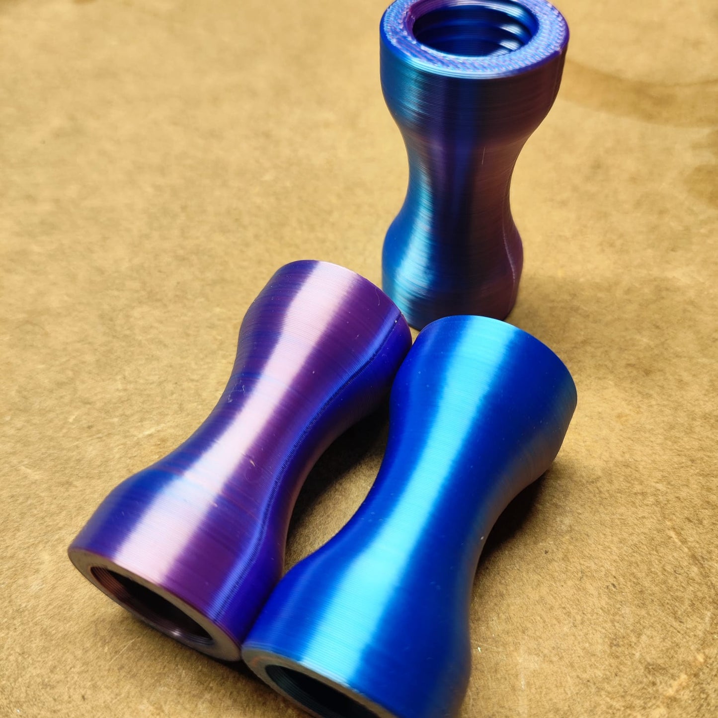 Drei metallische, sanduhrförmige Fahrrad-Steuersatz-Abstandshalter in Lila, Blau und Magenta liegen auf einer braunen Oberfläche. Der blaue Abstandshalter steht aufrecht wie das Schmales Griffstück für Modularen Diamond Painting Stift von New Plastics, während die violetten und magentafarbenen anmutig liegen.