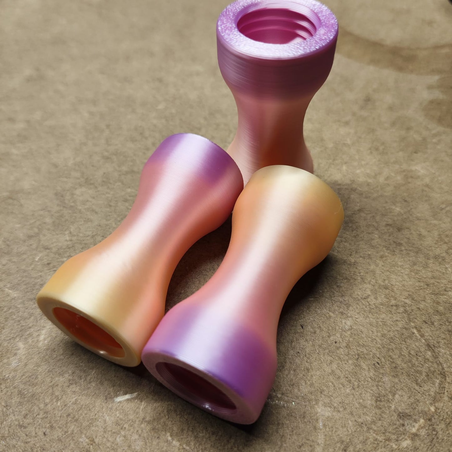 Drei farbenfrohe Metallobjekte in Sanduhrform weisen Farbverläufe in Rosa, Orange und Lila auf. Sie ähneln ergonomischen Formen und sind auf einer strukturierten braunen Oberfläche angeordnet – eines steht aufrecht wie ein Schmales Griffstück für Modularen Diamond Painting Stift, die anderen liegen auf der Seite.