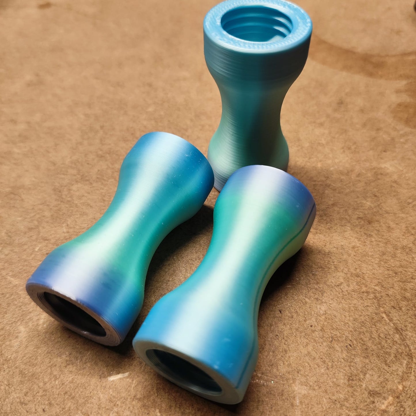 Drei kleine, ergonomische Objekte in Sanduhrform in Blau- und Grünverlauf liegen auf einer braunen Oberfläche, zwei liegen auf der Seite und eines aufrecht. Ihre glatte, glänzende Oberfläche erinnert an das Schmales Griffstück für Modularen Diamond Painting Stift von New Plastics.