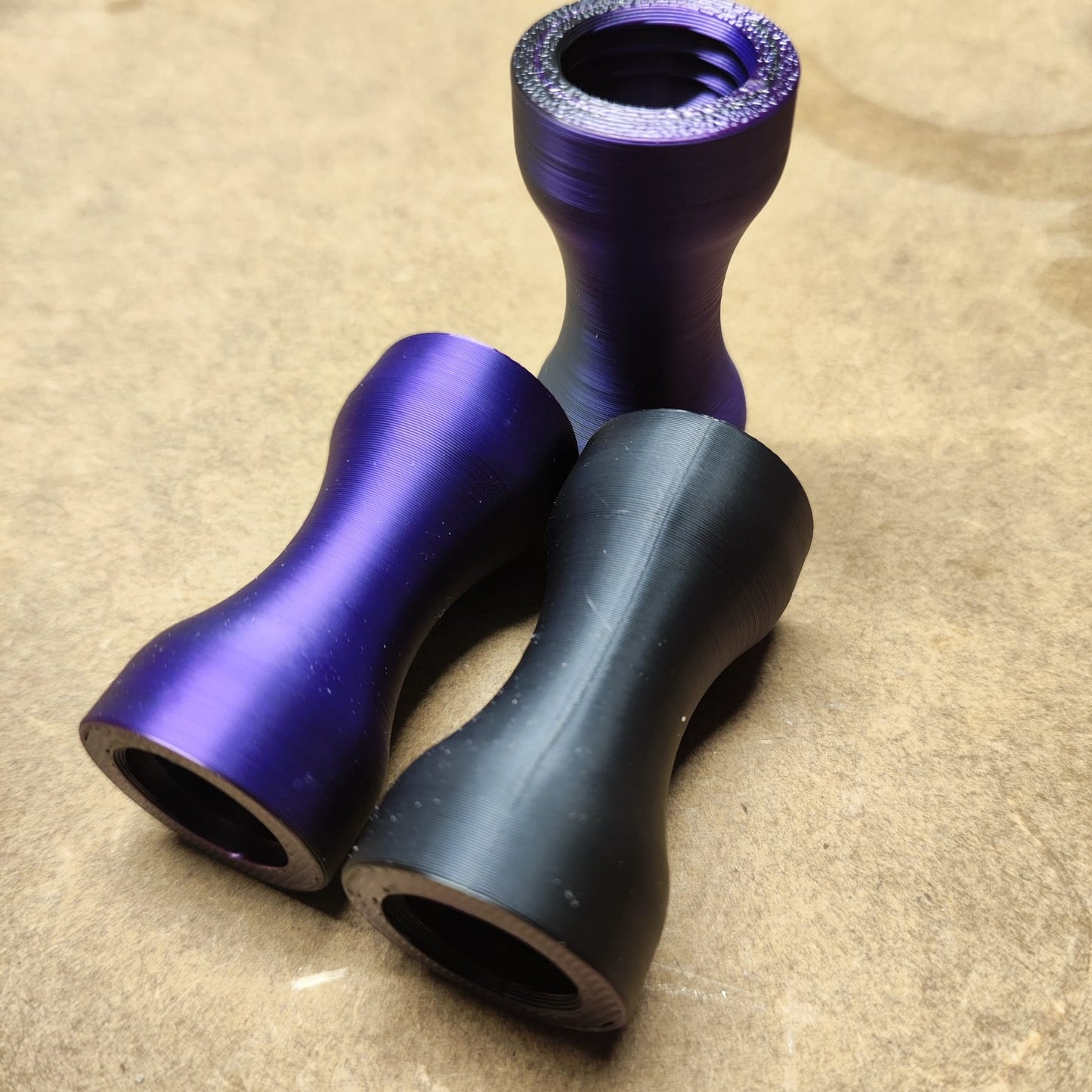 Drei eloxierte Metallteile, zwei violette und ein schwarzes, in Form des Schmales Griffstücks für den modularen Diamond Painting Stift von New Plastics, sind auf einer hellbraunen Oberfläche angeordnet. Sie zeichnen sich durch eine ergonomische Form mit glatter Textur und glänzender Oberfläche aus.