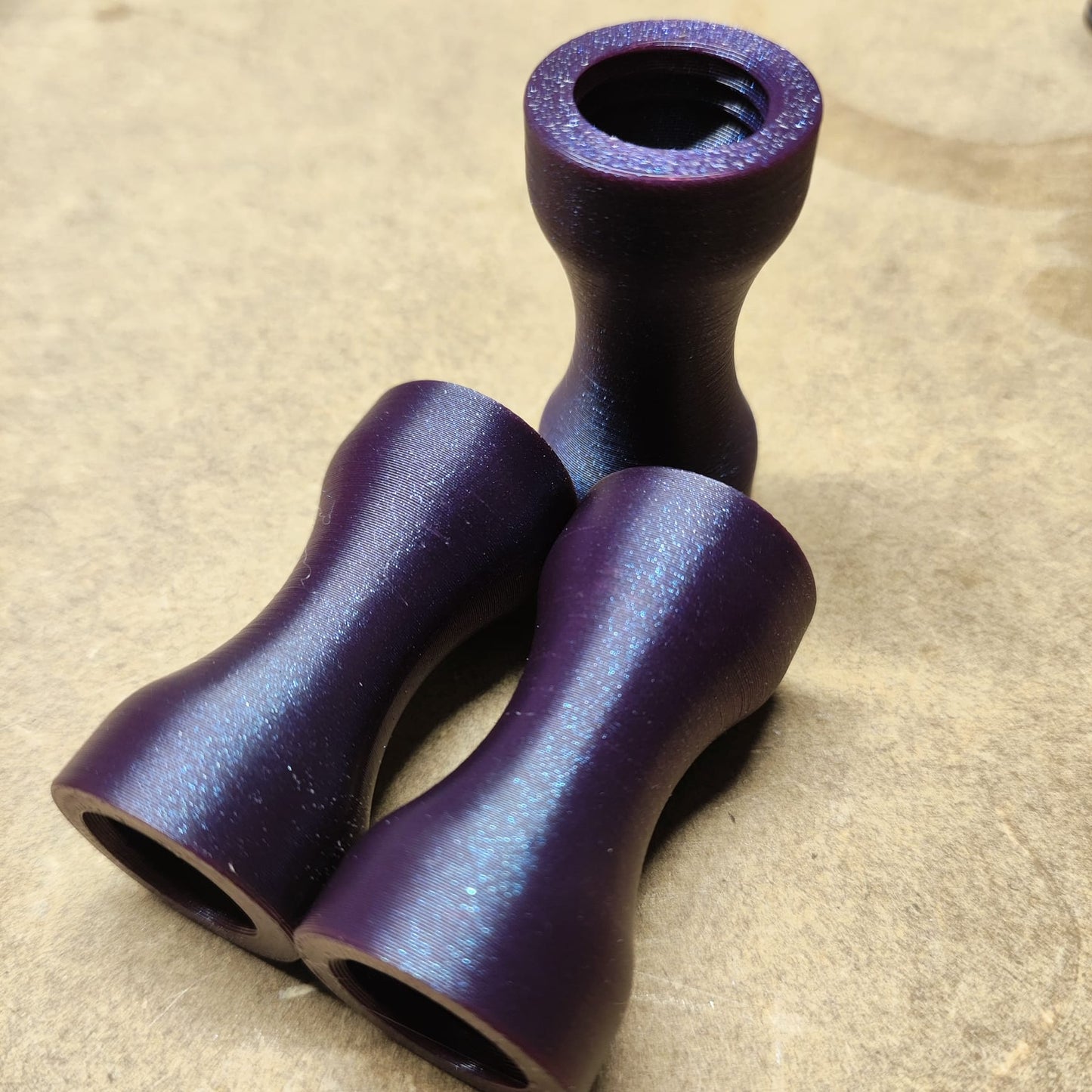 Drei lila, metallische Schmales Griffstücke von New Plastics liegen auf einer hellen Oberfläche. Mit ihrer ergonomischen Sanduhrform und der strukturierten Oberfläche sehen diese offenen Objekte wie kleine Vasen aus.