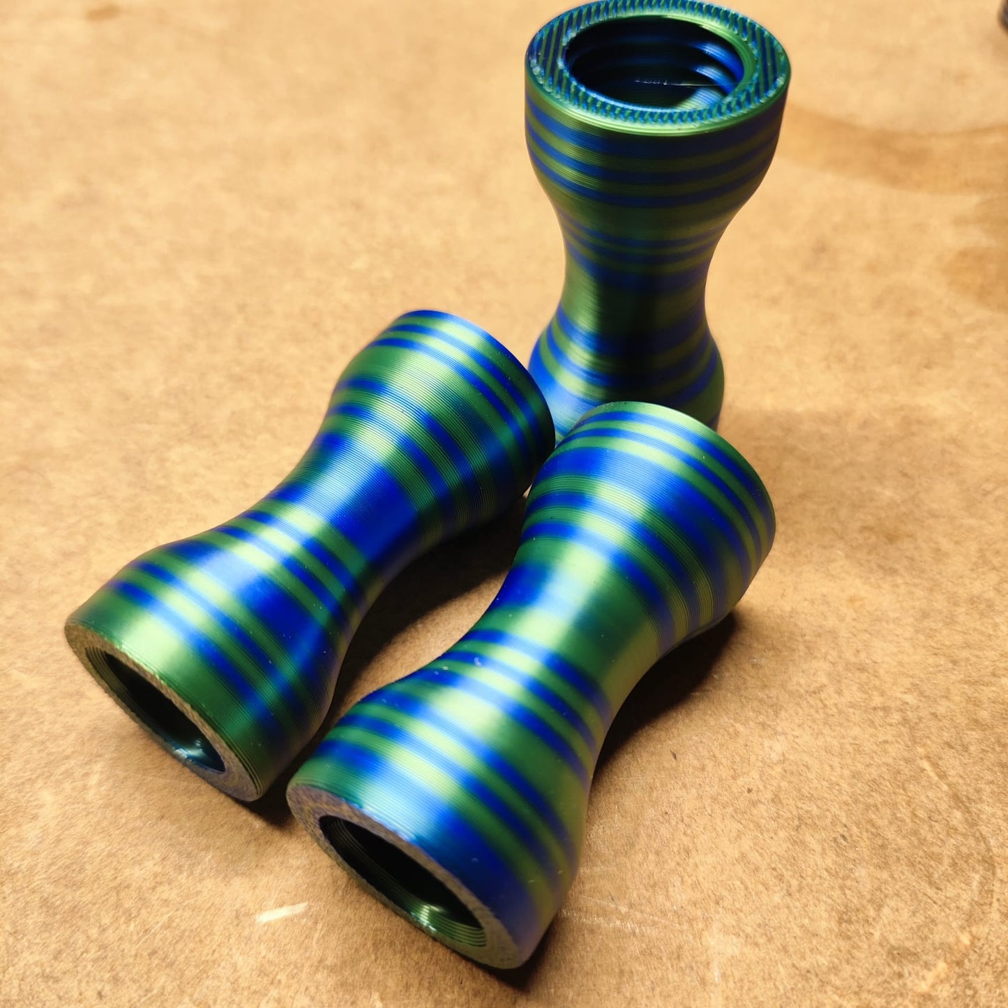 Drei metallische, sanduhrförmige Objekte mit blauen und grünen Streifen liegen auf einer braunen Fläche. Zwei liegen horizontal parallel zueinander, eines steht aufrecht. Sie ähneln dem ergonomischen Design des Schmales Griffstücks für Modularen Diamond Painting Stift von New Plastics.