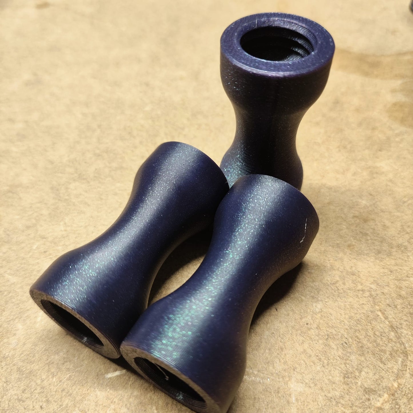 Drei dunkle Metallobjekte mit glattem, ergonomischem Design sind auf einer strukturierten beigen Oberfläche angeordnet. Unter dem Namen „Schmales Griffstück für Modularen Diamond Painting Stift *Abverkauf Nur solange der Vorrat reicht*“ werfen diese eleganten Artikel aus New Plastics weiche Schatten und reflektieren Licht, um ihre Kurven zu betonen.