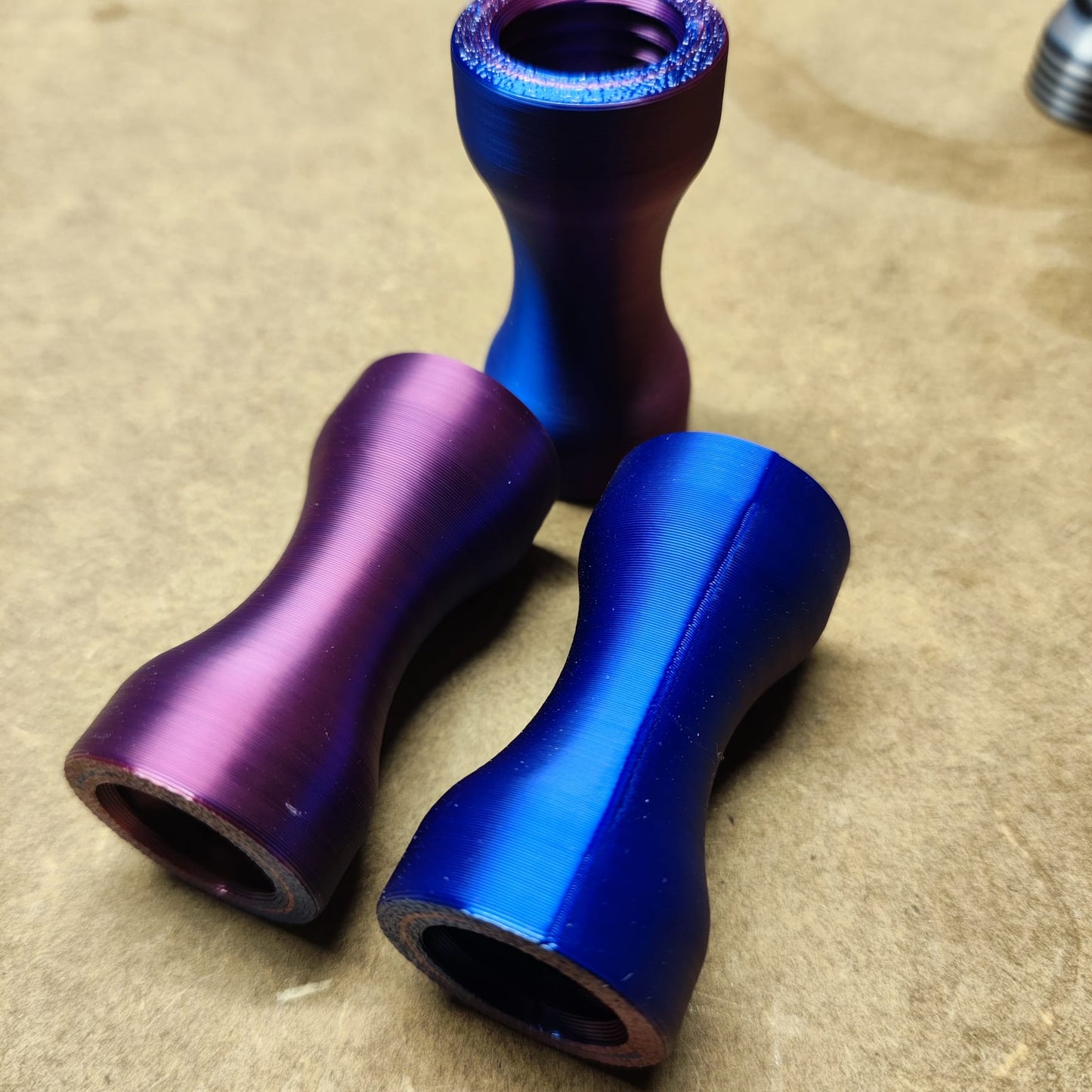 Drei Schmales Griffstück für Modularen Diamond Painting Stift von New Plastics, zwei in leuchtendem Lila und eines in tiefem Blau, zeigen eine ergonomische Sanduhrform auf beigem Untergrund. Ihre glänzende Oberfläche erinnert an dicht nebeneinander angeordnete Teile eines modularen Sets.