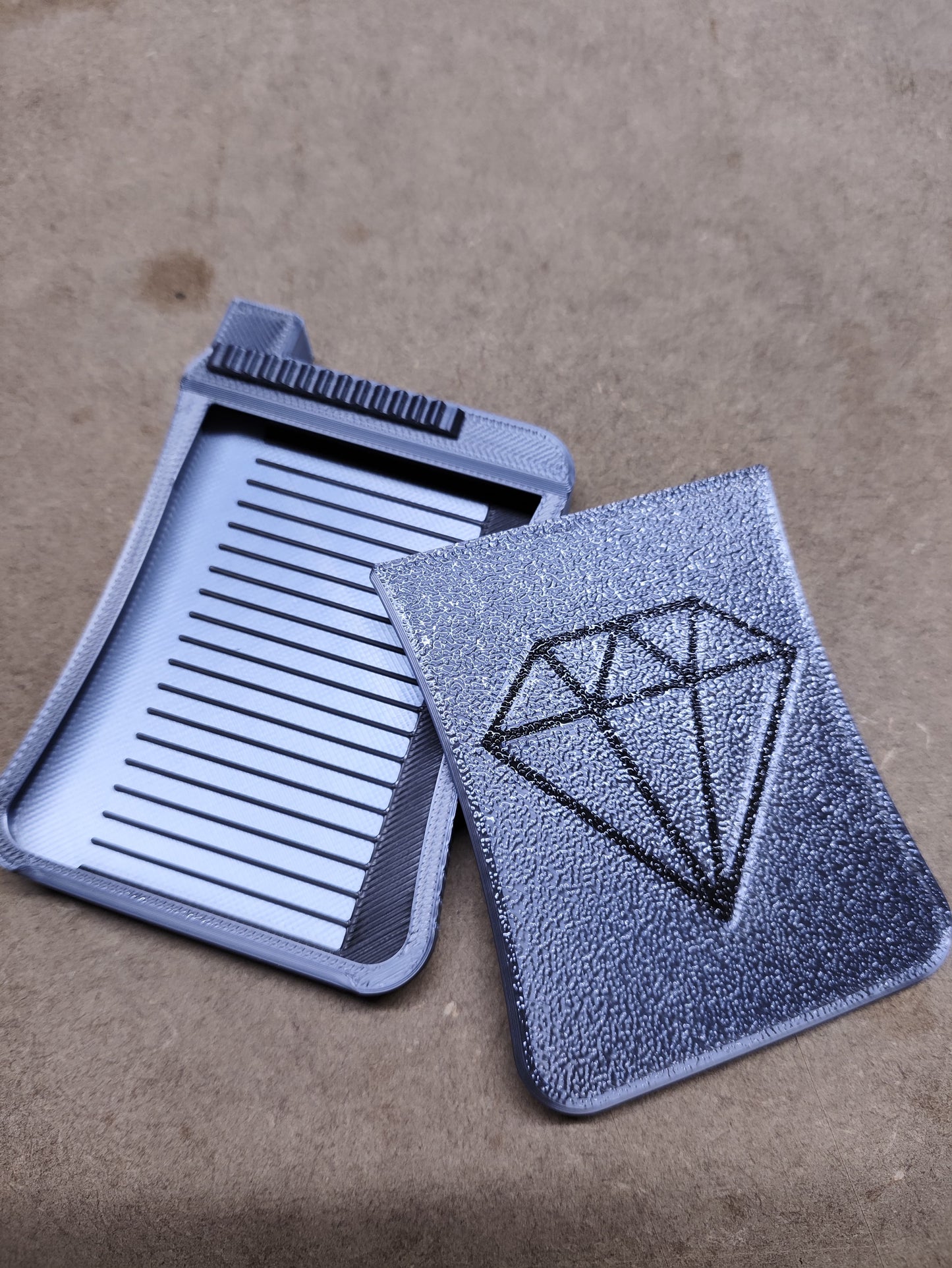 Zwei New Plastics Diamond Painting Schiffchen S3-Kartenhalter aus glattem silbernem und schwarzem Kunststoff liegen auf einer grauen Oberfläche. Einer ist geöffnet und zeigt sein charakteristisches geriffeltes Inneres; der andere bleibt geschlossen und weist ein markantes Diamantsymbol auf.