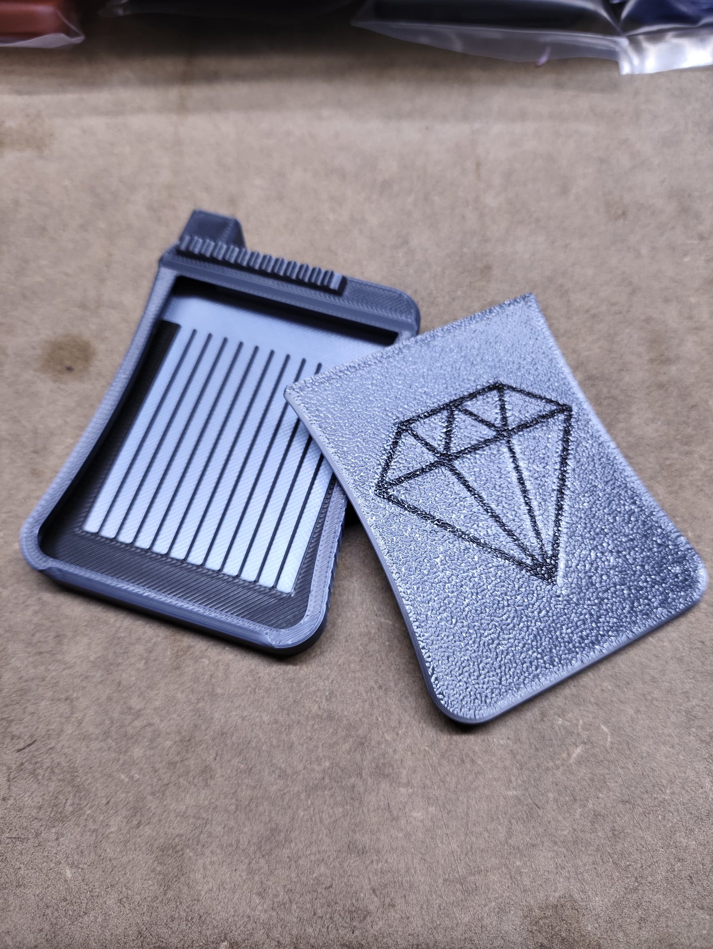 Ein grauer Kartenhalter „Diamond Painting Schiffchen S3“ von New Plastics liegt offen auf einer beigen Oberfläche und zeigt mehrere vertikale Kartenfächer und ein Rautenmuster.