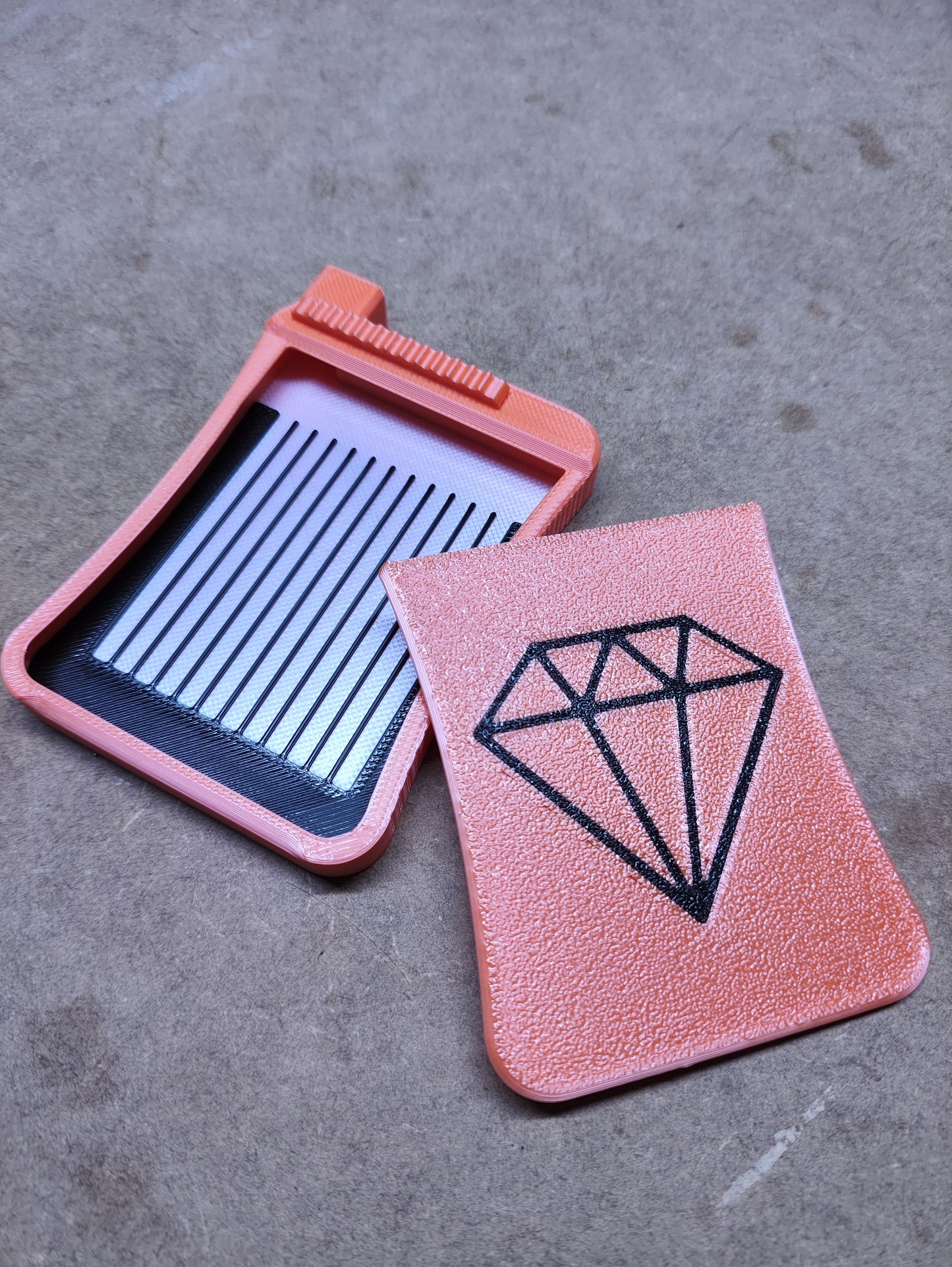 Auf einer grauen Oberfläche liegen zwei rosa Kunststoffwerkzeuge von New Plastics. Eines ist das Diamond Painting Schiffchen S3 mit schwarzem Rautendesign, das andere ist ein Haarschneider mit Metallzähnen und Griff. Beide Werkzeuge haben leicht strukturierte Oberflächen für Präzision.