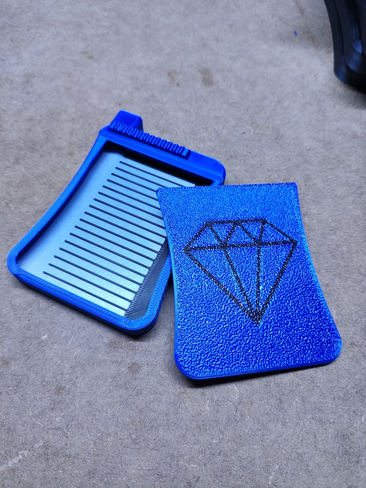 Auf einer strukturierten Oberfläche ruhen zwei blaue Kunststoffteile von New Plastics – ein rechteckiges Werkzeug mit horizontalen Rillen für Präzision im FDM-Verfahren und das Diamond Painting Schiffchen S3 mit Diamantgrafik, ideal für Enthusiasten.