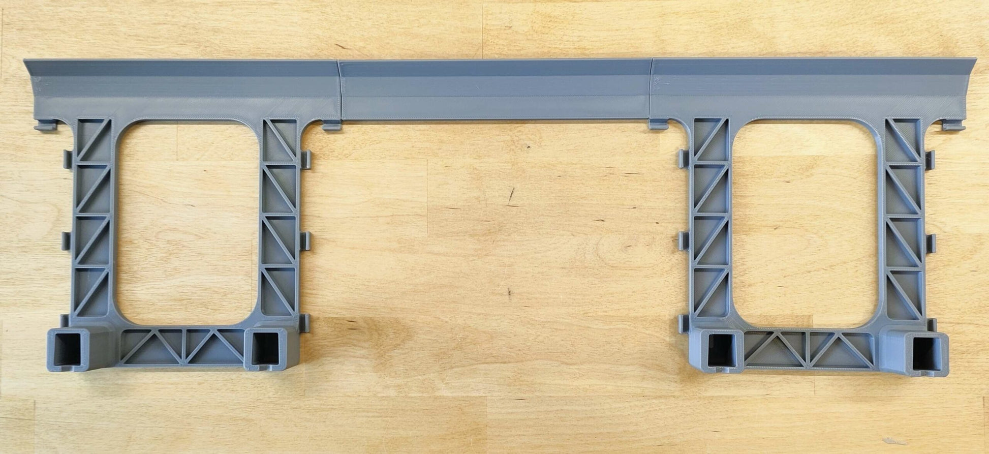 Eine graue LED-Beleuchtungshalterung von New Plastics für die Steinchen Garage, mit einer gitterartigen Struktur und zwei rechteckigen Öffnungen auf einer Holzoberfläche, bietet Montagebereiche an beiden Enden, ideal für die Organisation Ihres LED-Streifen-Setups.