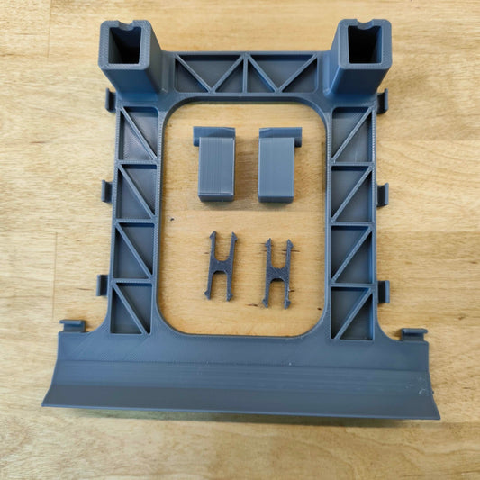 Ein graues 3D-gedrucktes Objekt von New Plastics, das einer LED-Beleuchtungshalterung für Steinchen Garage ähnelt, liegt auf einer Holzoberfläche. Es besteht aus einem quadratischen Rahmen mit sich kreuzenden Stützen, der zwei rechteckige Teile und zwei kleinere „H“-förmige Komponenten innerhalb des Rahmens enthält.