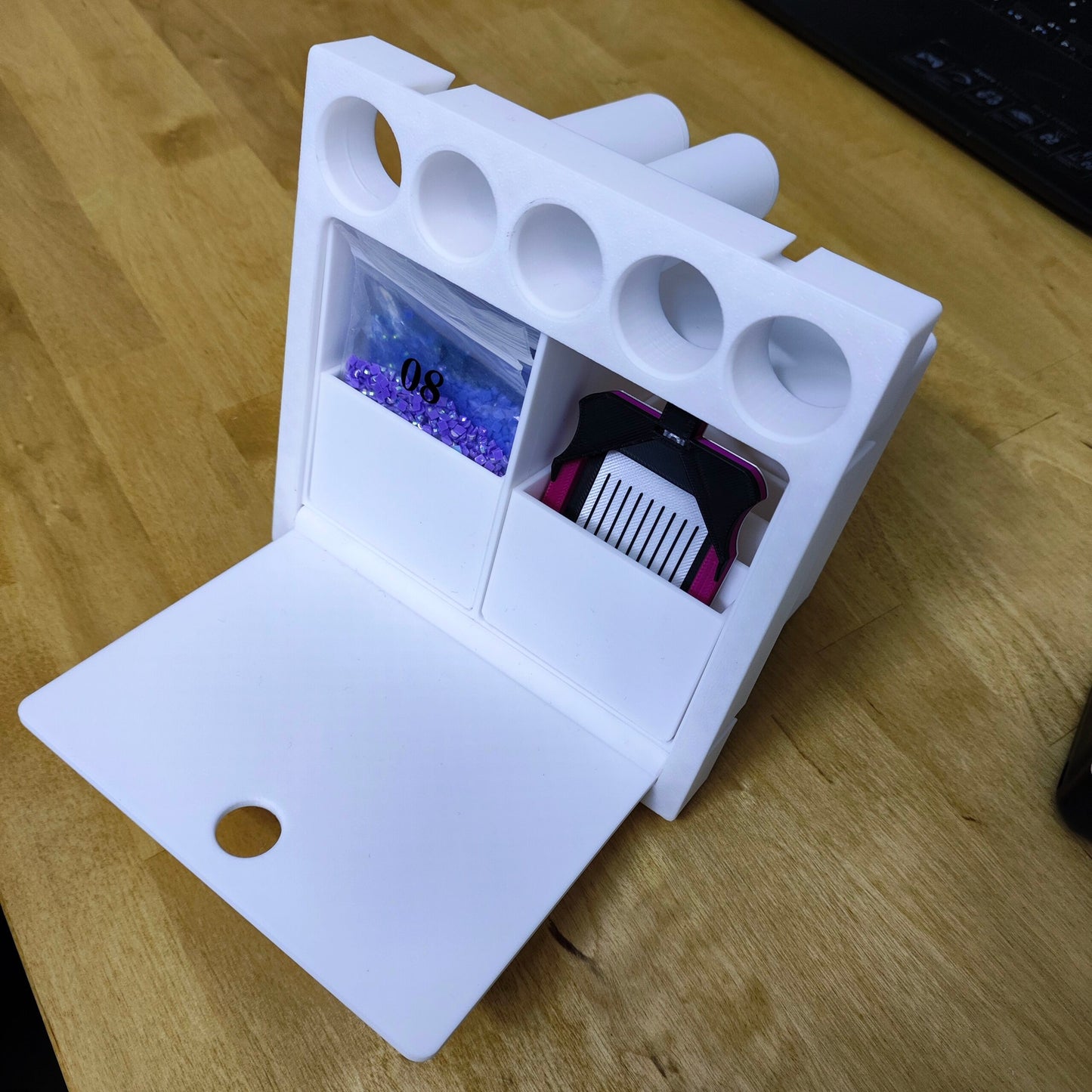 Auf einer Holzoberfläche hält ein weißes Multitalent-Modul für Steinchen Garage von New Plastics kleine violette Perlen und einen schwarz-rosa Gegenstand. Es hat oben zylindrische Schlitze und vorne eine geschlossene Klappe, was es zu einem idealen Bastelmodul mit Schubfach macht.