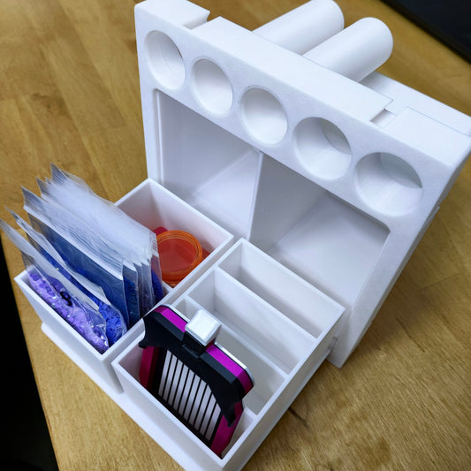 Das New Plastics Multitalent Modul für Steinchen Garage, ein 3D-gedruckter Organizer in Weiß, steht auf einer Holzoberfläche. Es enthält mehrere Diamondpainting-Tütchen, einen runden Behälter und einen gestreiften Gegenstand mit Clip. Die Oberseite hat Löcher für stiftähnliche Gegenstände.