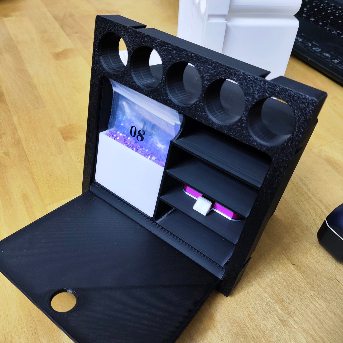 Das New Plastics Multitalent Modul für Steinchen Garage, ein Organizer aus schwarzem Kunststoff auf Holzoberfläche, verfügt oben über vier runde Abschnitte und darunter über mehrere Fächer. Eine kleine Kalenderseite zeigt „08“ und die sichtbaren Schubladen sind ideal zum Ordnen von Diamondpainting-Tütchen oder Zubehör.