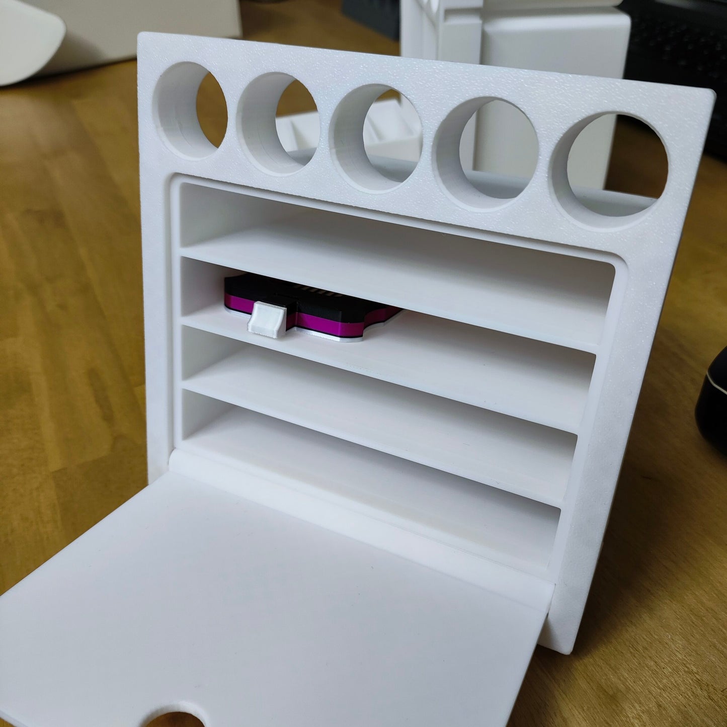 Das weiße 3D-gedruckte New Plastics Multitalent Modul für Steinchen Garage hat oben fünf runde Fächer und unten vier horizontale Schlitze, darunter einen für ein kleines rosa USB-Gerät. Dieses Design, das einem Modul mit Schubfach ähnelt, wird elegant auf einer Holzoberfläche präsentiert.