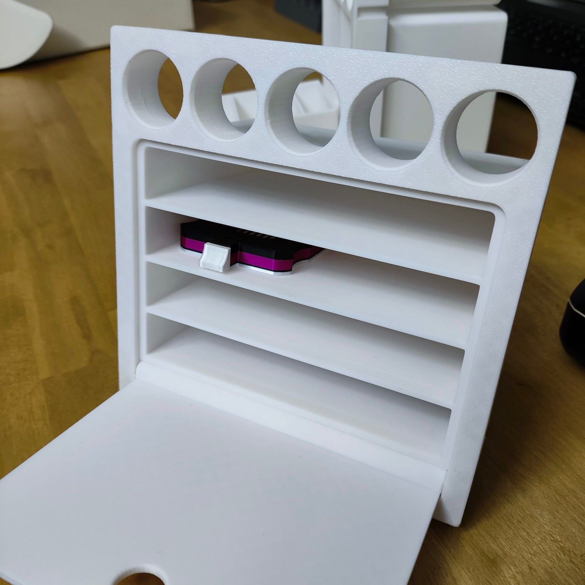 Das weiße 3D-gedruckte New Plastics Multitalent Modul für Steinchen Garage hat oben fünf runde Fächer und unten vier horizontale Schlitze, darunter einen für ein kleines rosa USB-Gerät. Dieses Design, das einem Modul mit Schubfach ähnelt, wird elegant auf einer Holzoberfläche präsentiert.