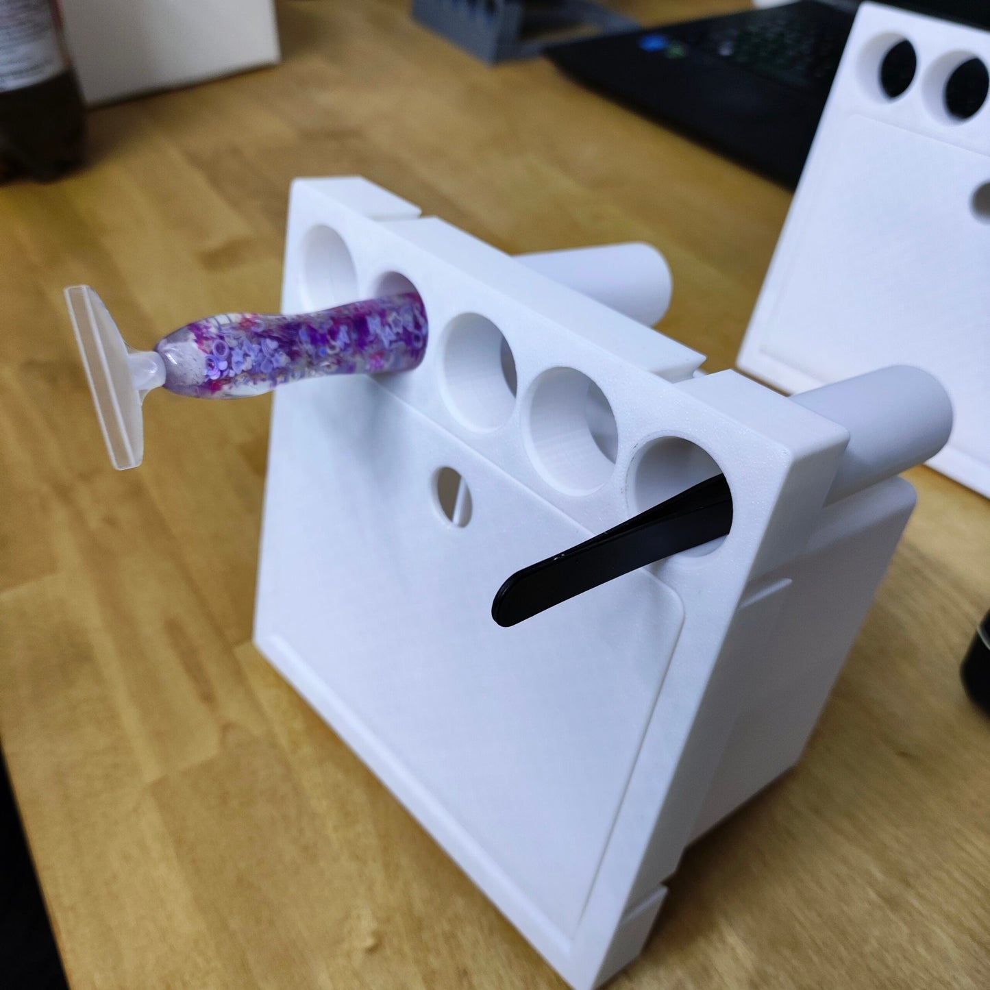 Eine Nahaufnahme eines weißen 3D-gedruckten „Stifte und Pinzetten Einsatz für Steinchen Garage“ von New Plastics auf einem Holzschreibtisch enthält eine rosa Zahnbürste und einen schwarzen Gegenstand. Zusätzliche Löcher könnten für Diamond Painting Stifte geeignet sein. Ein verschwommener Laptop und eine Flasche bilden im Hintergrund ein künstlerisches Tableau.