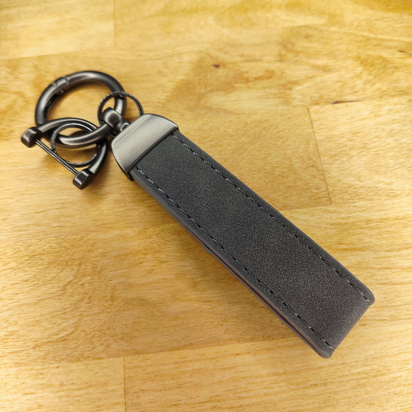 Eine Nahaufnahme des Schlüsselanhängers Leder Personalisiert von New Plastics mit grauem Riemen und Metallbeschlägen, der auf einer Holzoberfläche liegt.