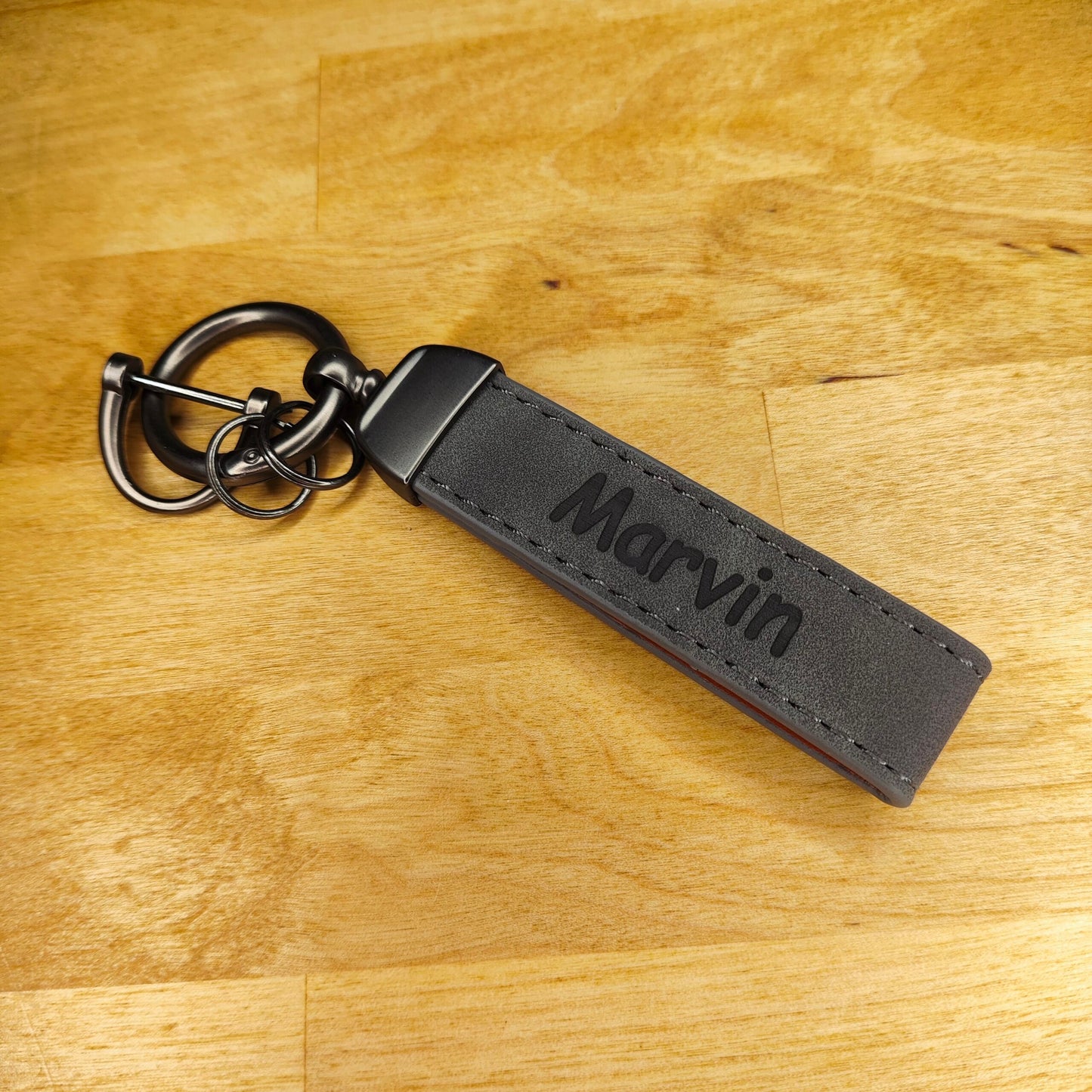 Der Schlüsselanhänger Leder Personalisiert von New Plastics mit der Prägung „Marvin“ liegt auf einer Holzoberfläche. Mit einem Metallring und einem Verschluss verbindet er Eleganz und Funktionalität perfekt.