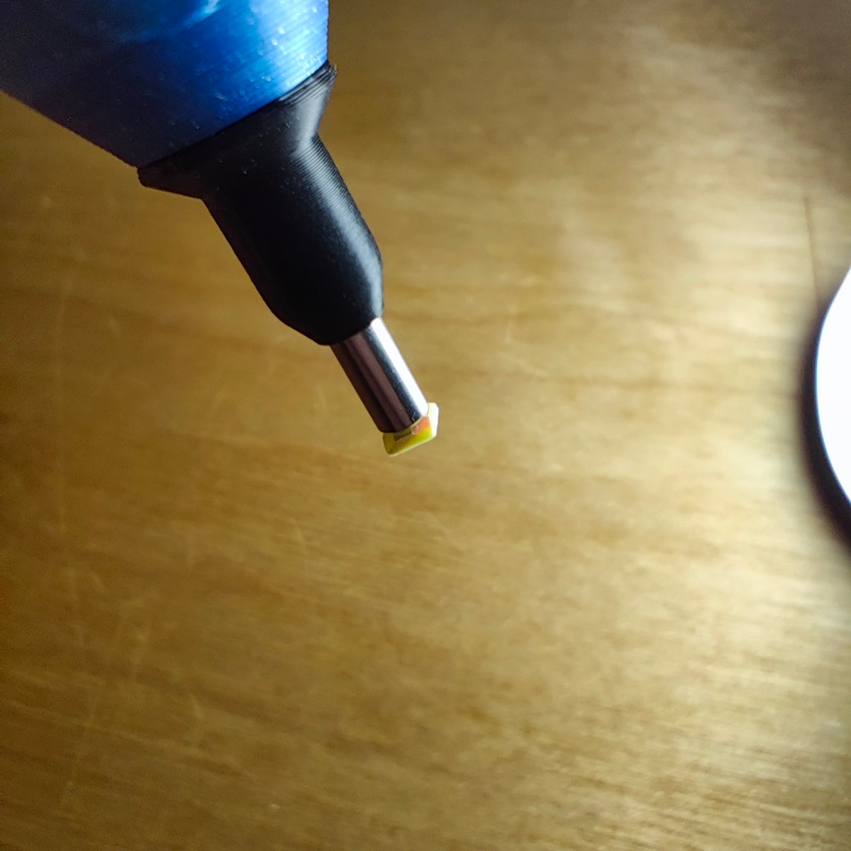 Nahaufnahme des New Plastics Diamond Painting Edelstahl Einzelsetzers V3, ein stiftähnliches Werkzeug mit einer Metallspitze, das ein kleines durchscheinendes gelbes Stück hält, vor einem strukturierten hellen Holzhintergrund, das Spitzenleistung demonstriert.