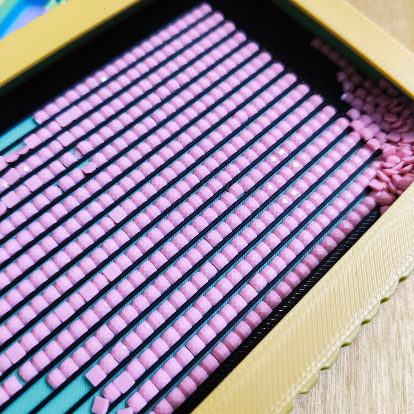 Eine Nahaufnahme eines Diamond Painting Schiffchen S1 PLUS von New Plastics zeigt eckige rosa Steinchen, die sauber auf einer schwarzen Klebeleinwand in einem gelben Rahmen angebracht sind, mit losen Steinen und einem Schiffchen in der Nähe, die darauf warten, dass sich ihre Kunstfertigkeit entfaltet.