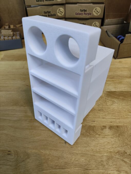 Ein weißes 3D-gedrucktes Objekt von New Plastics, hergestellt im FDM-Verfahren, hat oben zwei runde Öffnungen und darunter mehrere rechteckige Schlitze. Es steht auf einer Holzoberfläche mit Kartons im Hintergrund. Produkt: Mini Garage für 3 Schiffchen NP-S1, 4 Pinzetten und 1 Nahttrenner für Steinchen Garage.