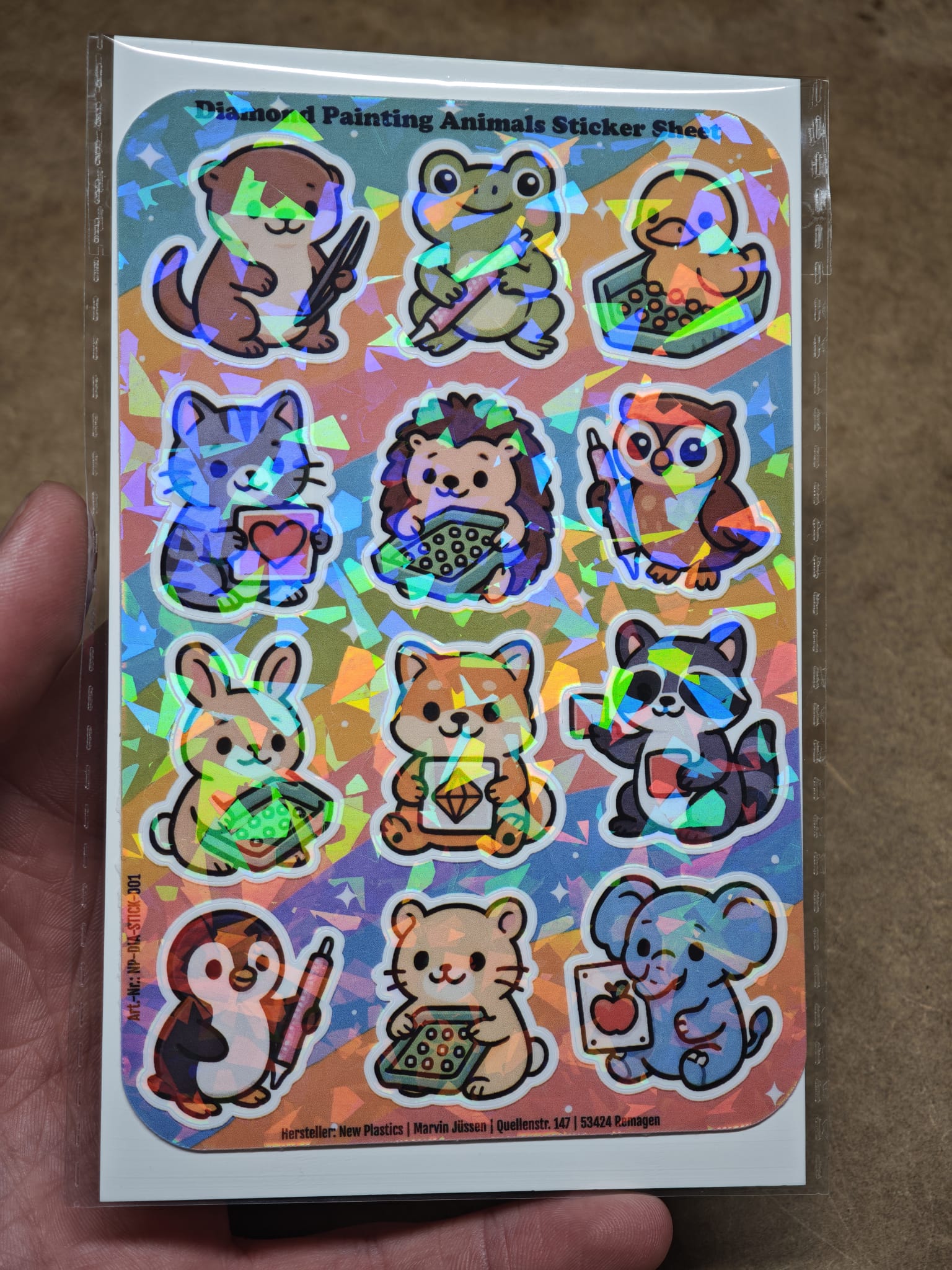 Eine Hand hält den New Plastics Diamond Painting Animals Sticker Sheet mit 12 Stickern ca. DIN A6, mit Cartoon-Tieren wie Hund, Frosch, Katze, Bär und mehr auf buntem Hintergrund – perfekt für DIY-Projekte mit Diamond Painting Werkzeugen.