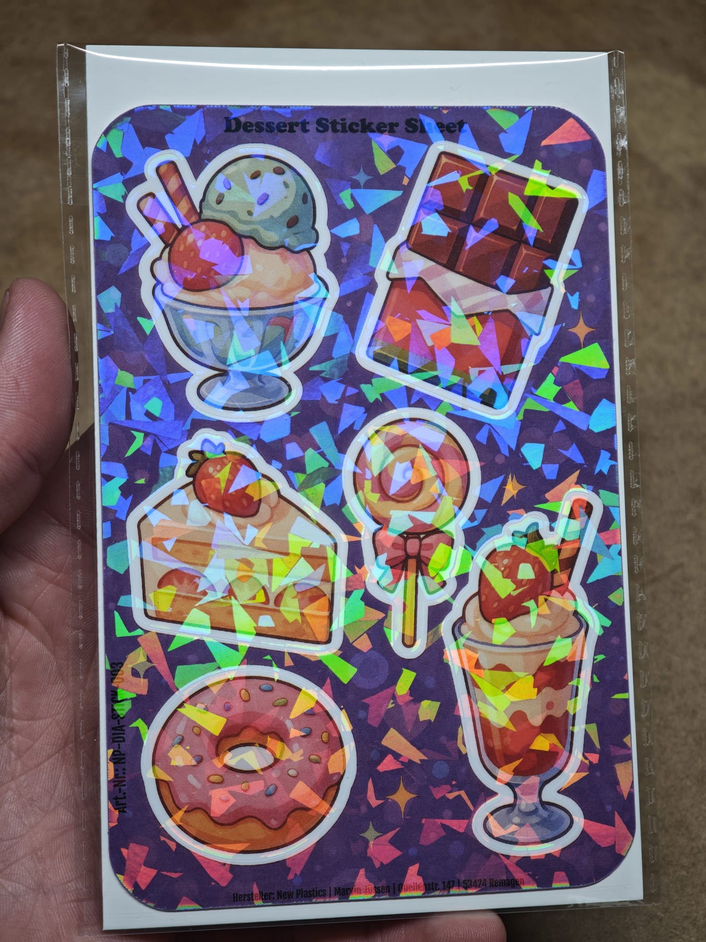 Eine Hand hält den New Plastics Dessert Sticker Sheet mit 6 Stickern ca. DIN A6 – mit Eis, Schokolade, Kuchen, Lutscher, Donut und Parfait – für DIY-Projekte vor einem leuchtend violetten Hintergrund.